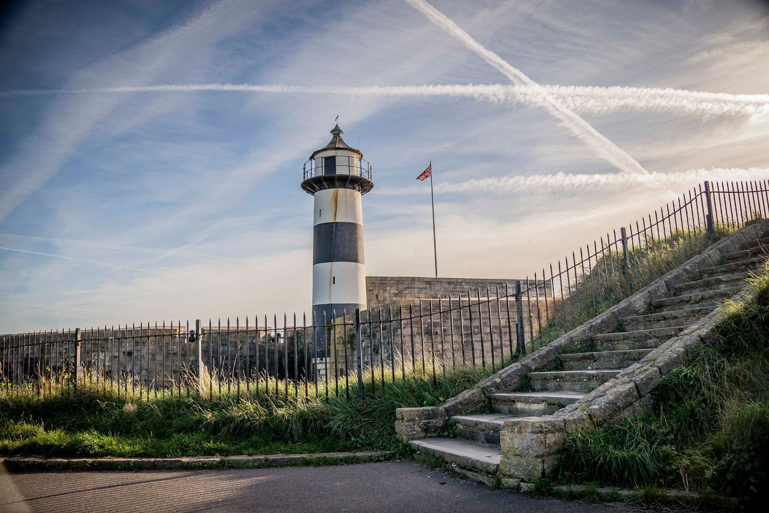 Coastal lighthouse and fence silhouetted against bright daytime sky.