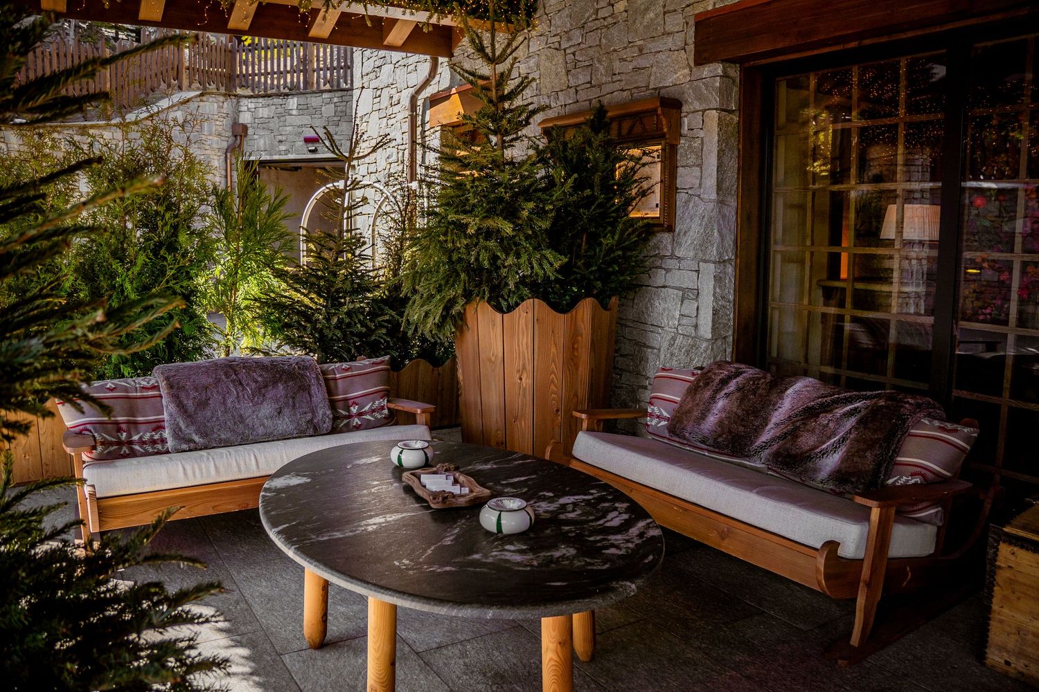 Cozy outdoor patio seating area with wooden benches, purple cushions, and potted plants against a stone wall.