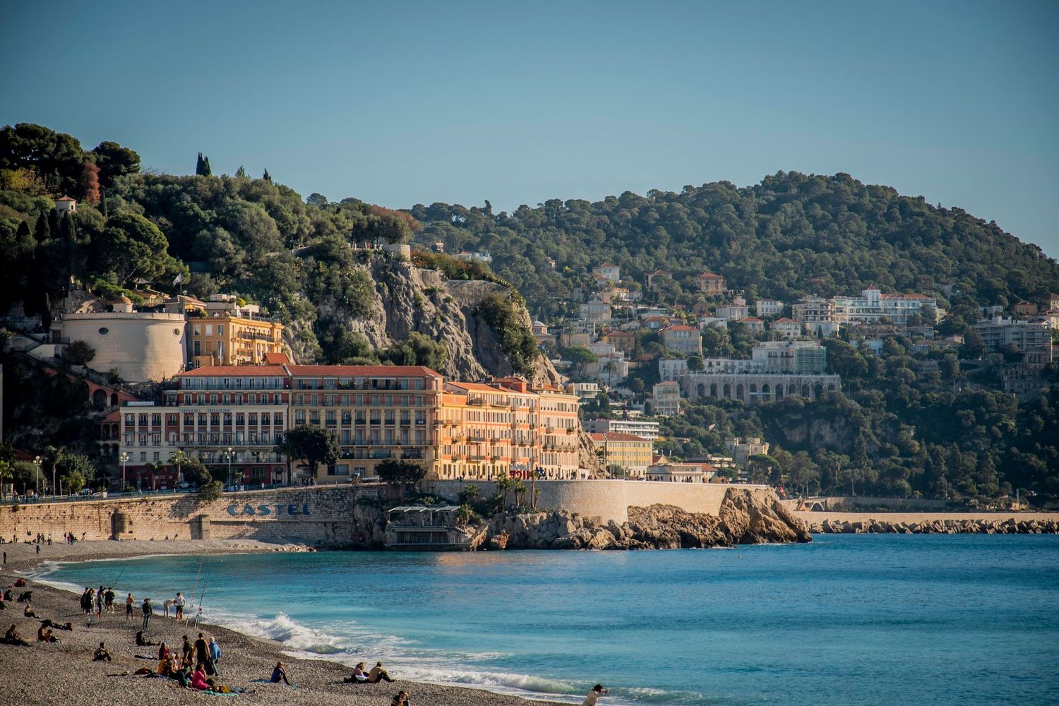 Coastal Mediterranean beach with historic beachfront buildings nestled against hills in Nice, France during golden hour.