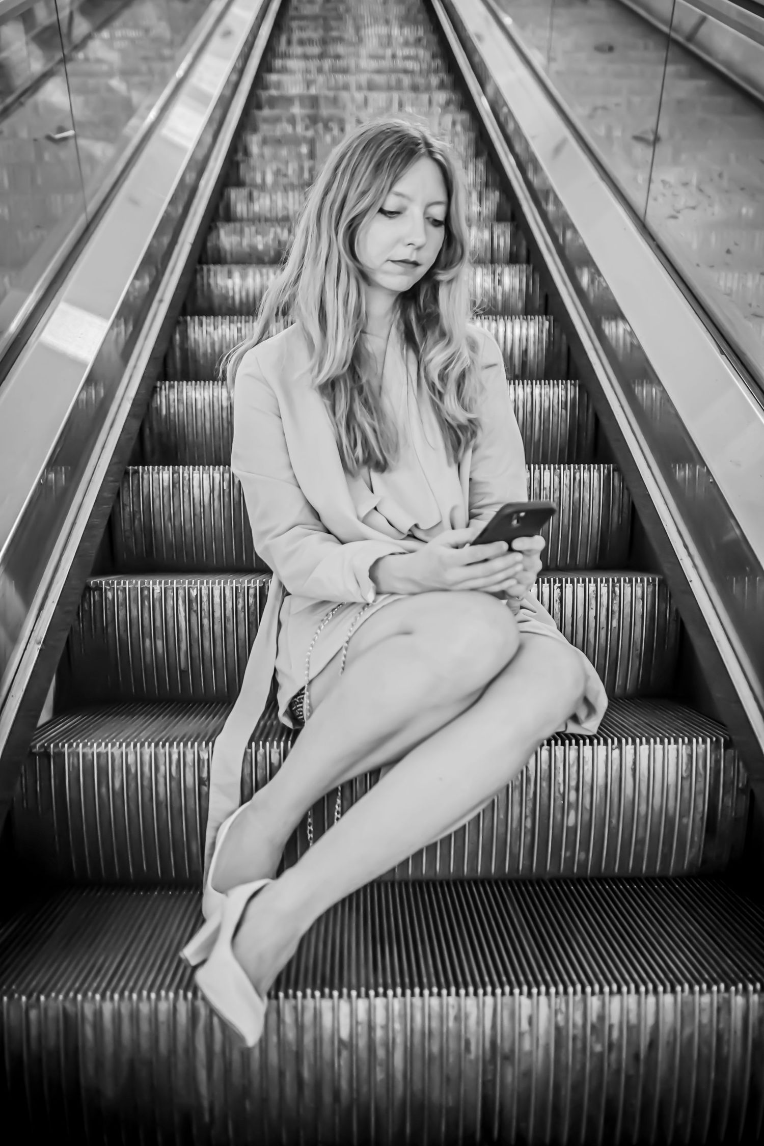 Woman in beige coat sitting on escalator steps checking phone in black and white.