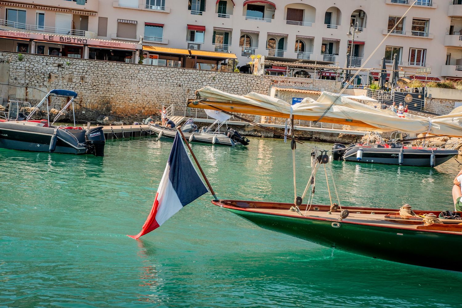 Marina with boats docked along a waterfront promenade featuring turquoise water and coastal buildings.