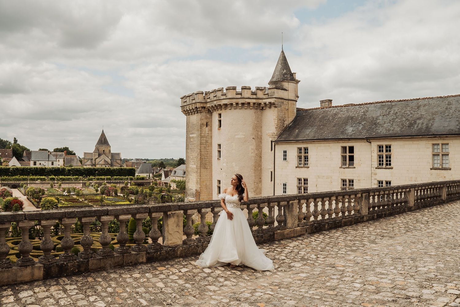 A romantic wedding couple walks along the stone ramparts of a historic French chateau under cloudy skies.