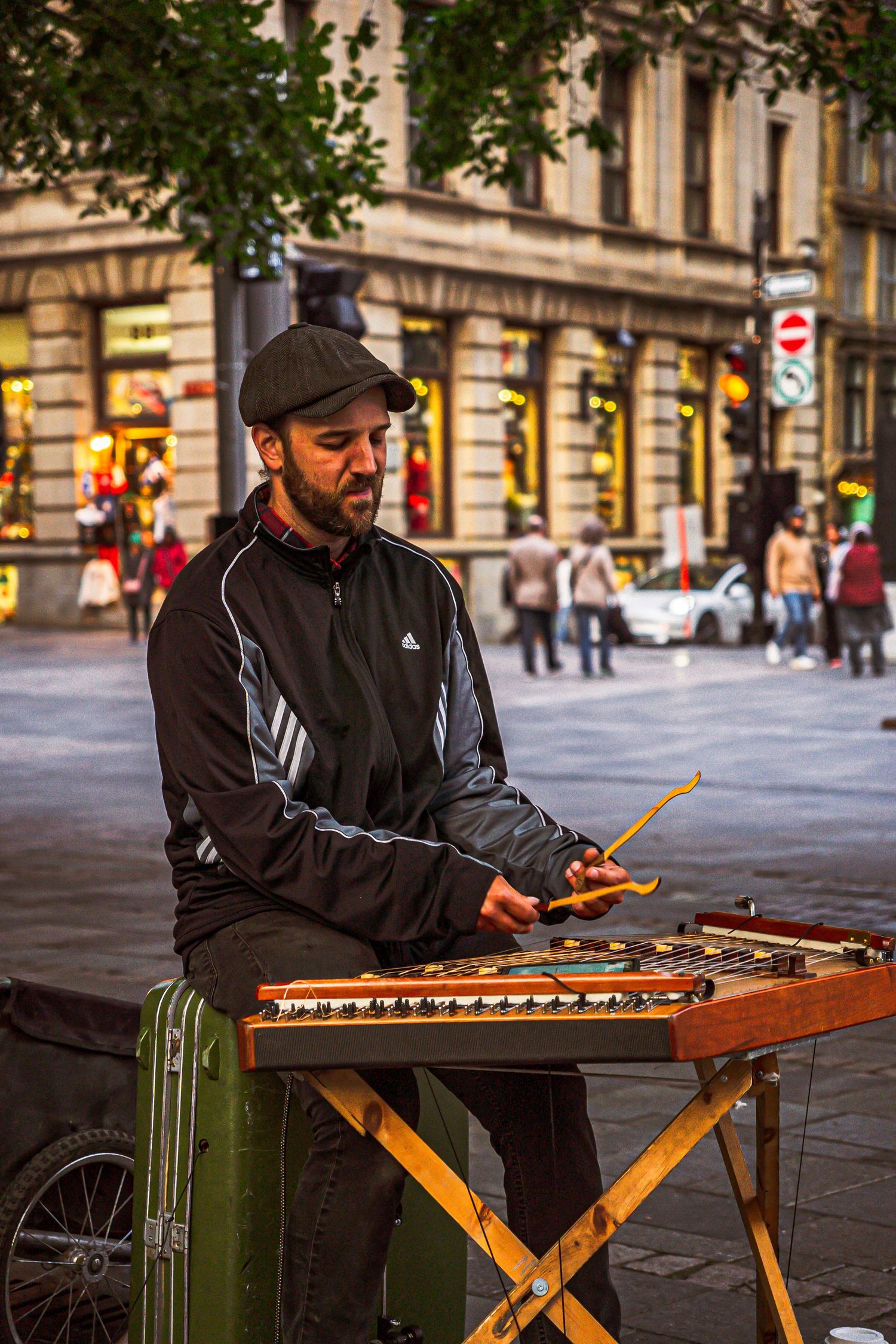 Street musician plays a unique wooden stringed instrument on a city sidewalk outside historic buildings.