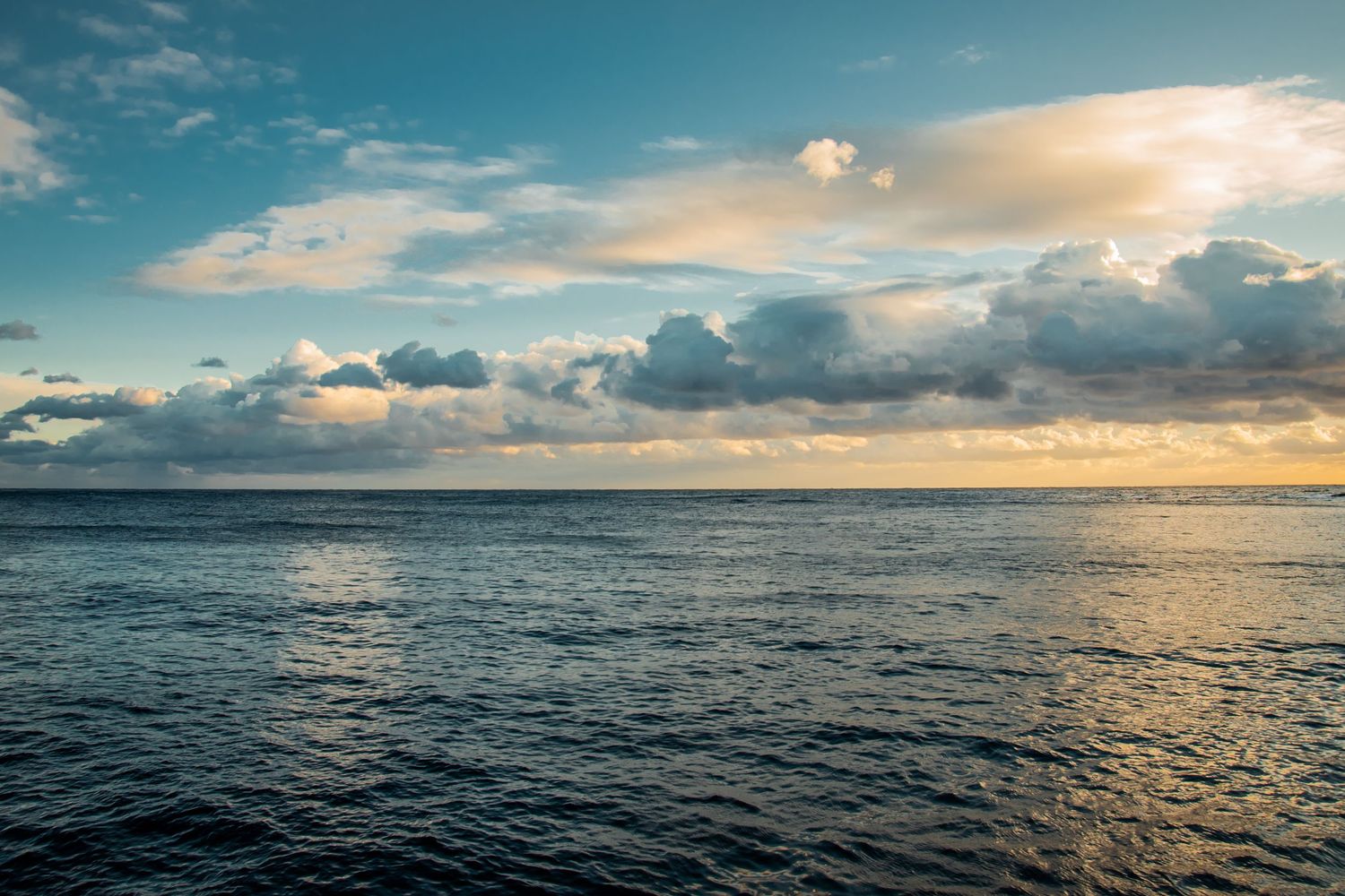 Dramatic sunset over a vast ocean horizon with scattered clouds reflecting golden light on rippling blue waters.