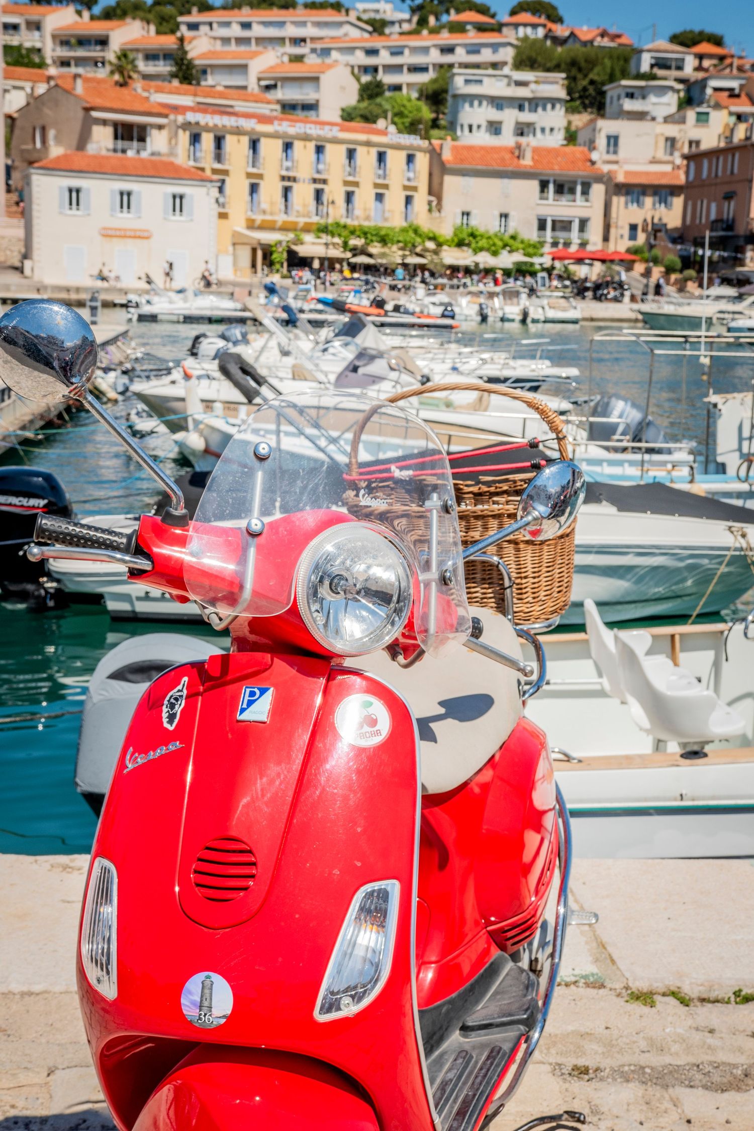 Red Vespa scooter parked at Mediterranean marina with boats and coastal buildings in background.
