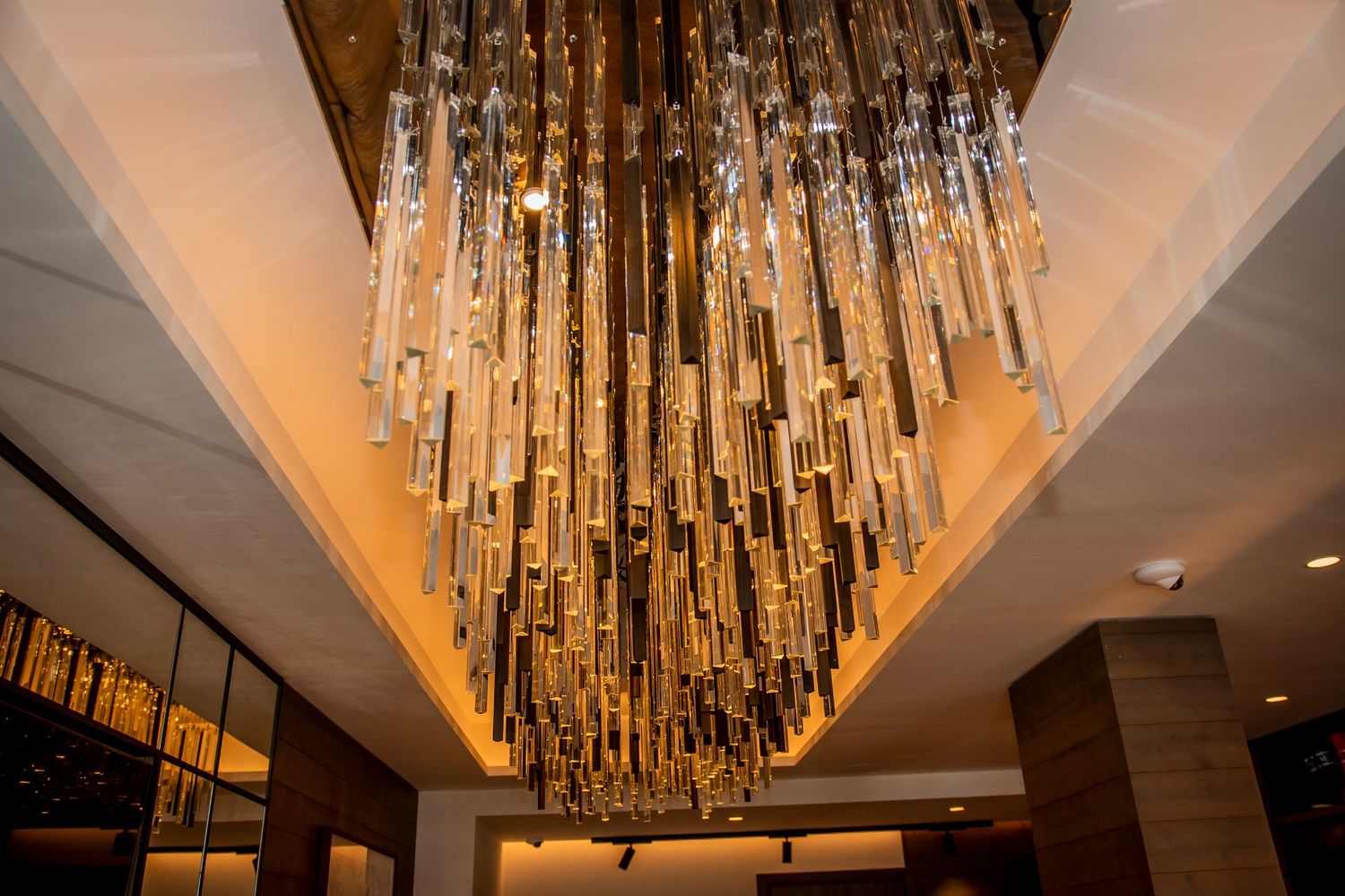 A luxurious modern chandelier with cascading glass tubes illuminates a hotel lobby ceiling with warm amber lighting.