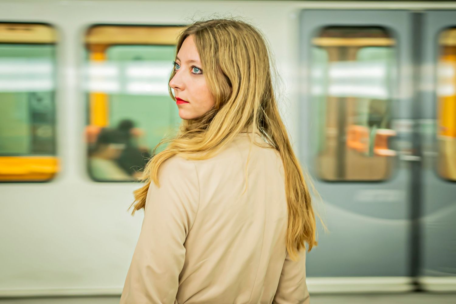 Woman in beige coat standing on subway platform with train car in background.