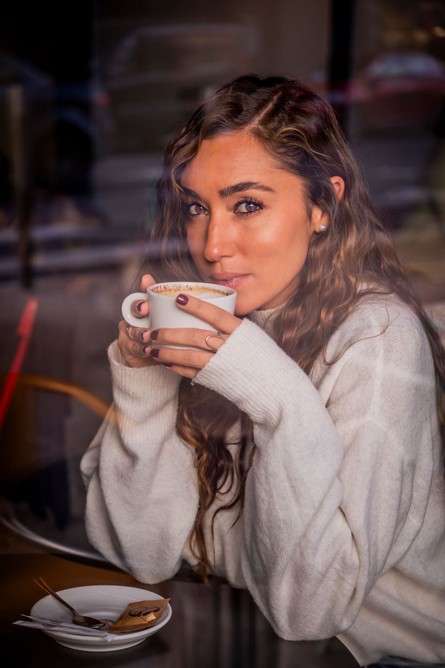 Person in cozy sweater drinks from white cup in warm indoor lighting.