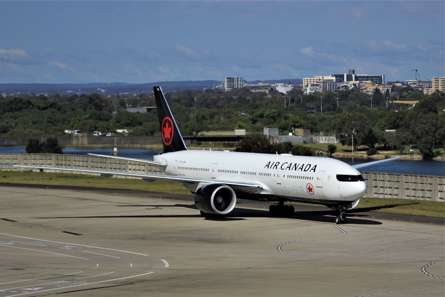 Air Canada Boeing 777 aircraft parked on tarmac with city buildings visible in background on sunny day.