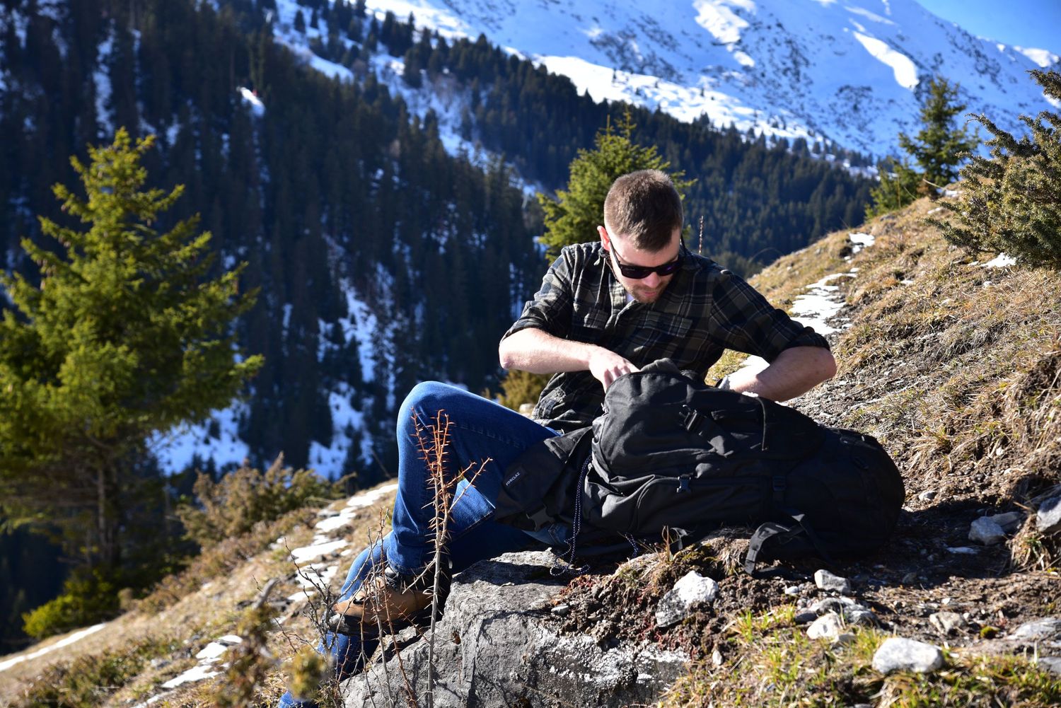 Hikers rest on rocky mountain overlook with snowy peaks and evergreen forests in scenic alpine landscape.