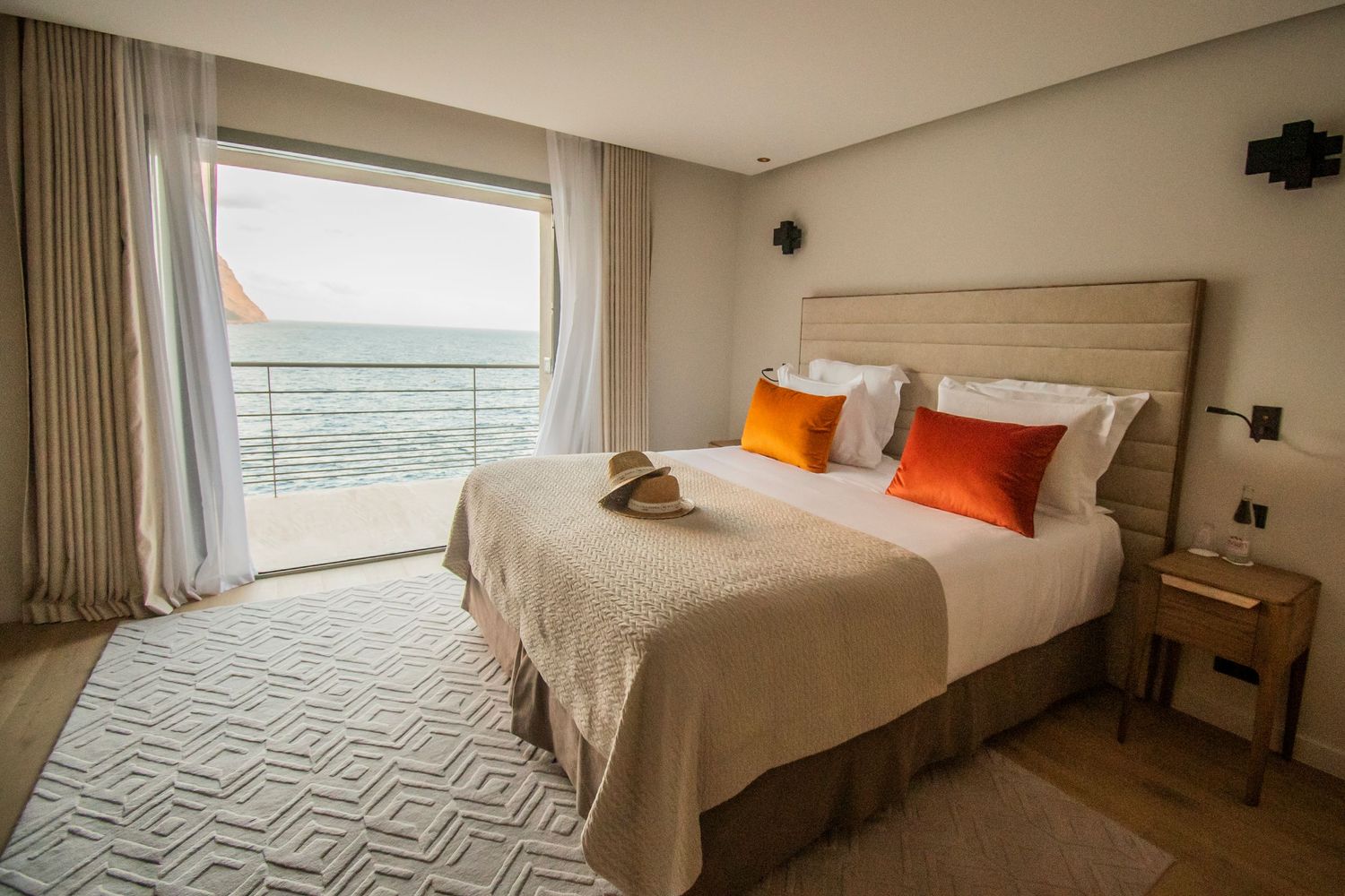Modern minimalist hotel room interior with white bedding and coastal view through large windows.