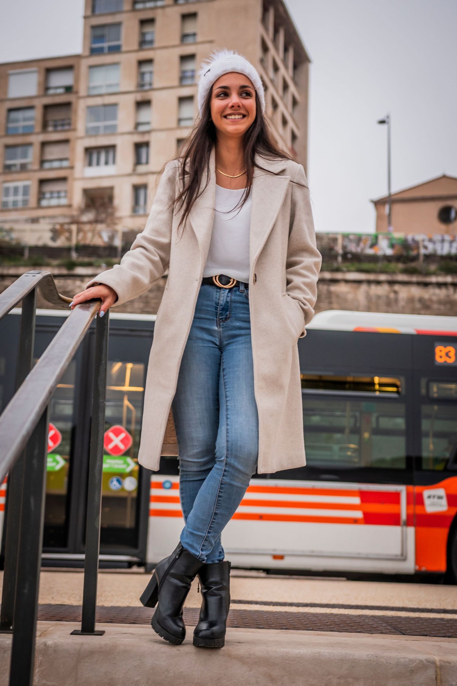 A person in winter fashion poses by a city bus stop wearing a beige coat and knit hat.