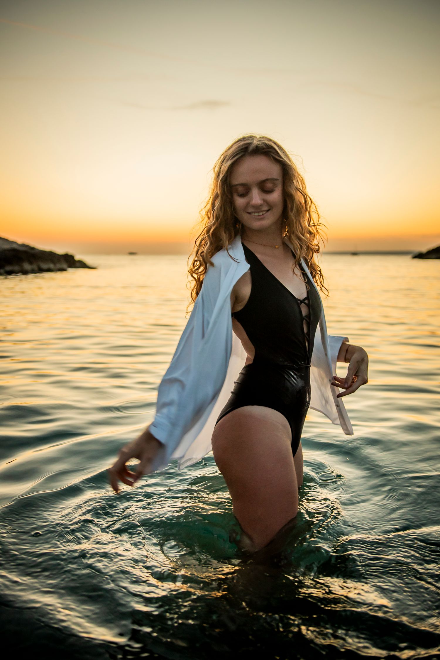 Woman wading in ocean water at sunset wearing black swimsuit and white shirt.