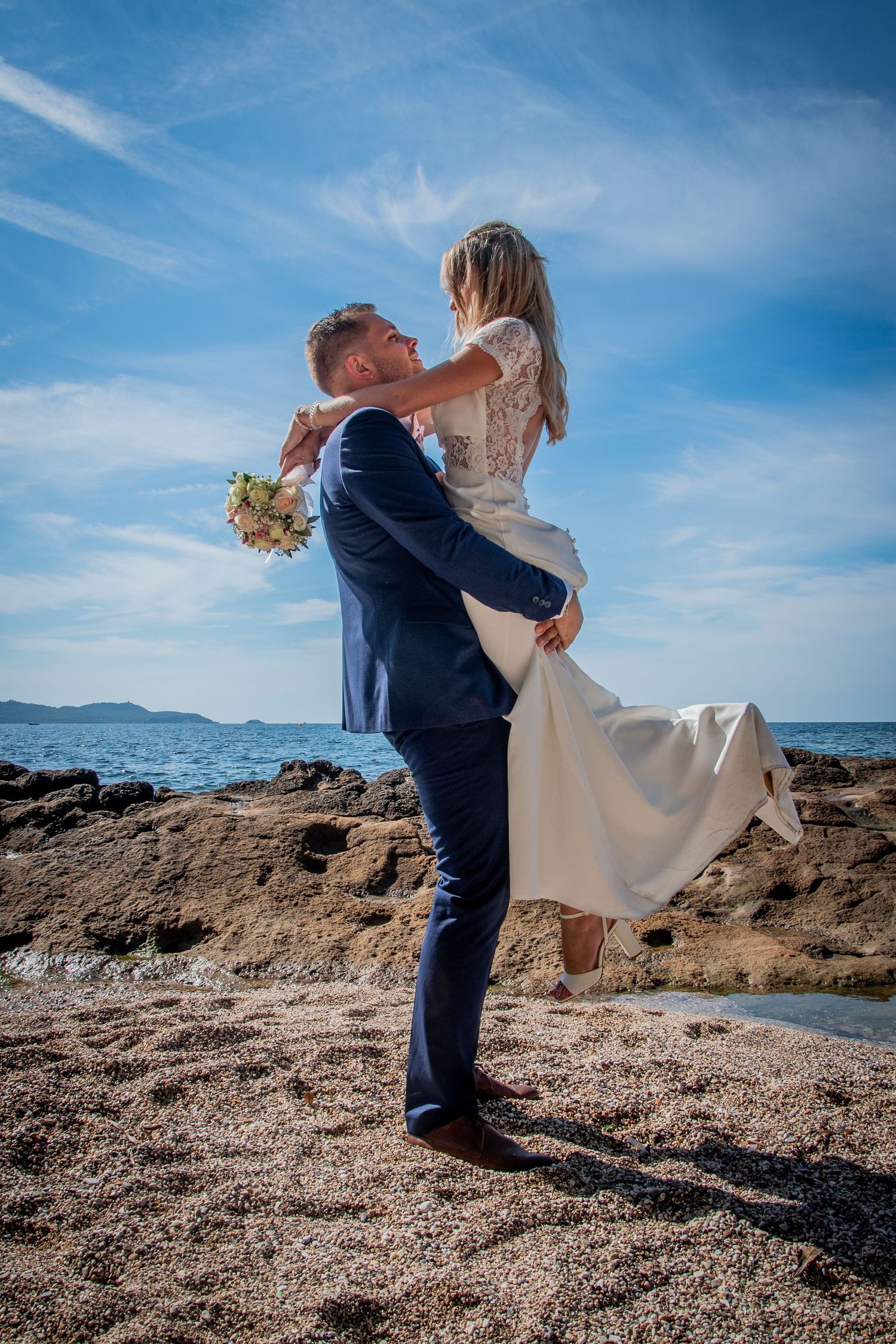 Coastal wedding photoshoot on rocky beach with blue ocean backdrop.