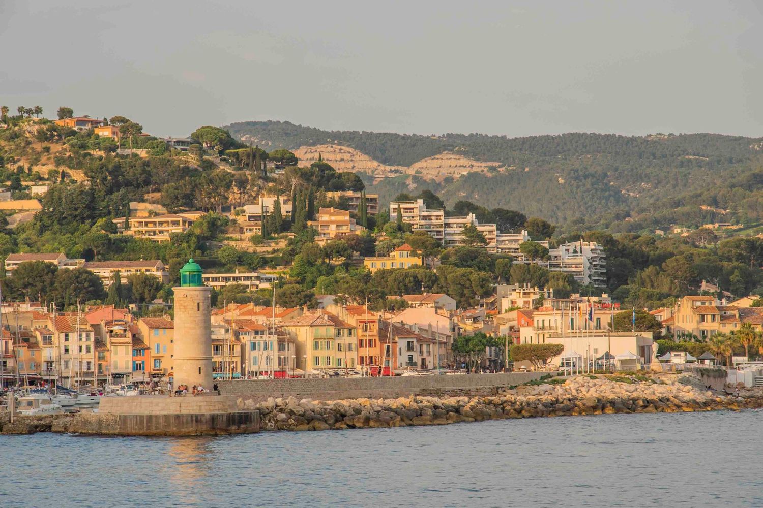 Mediterranean coastal town with colorful buildings and lighthouse nestled against hillside at sunset.