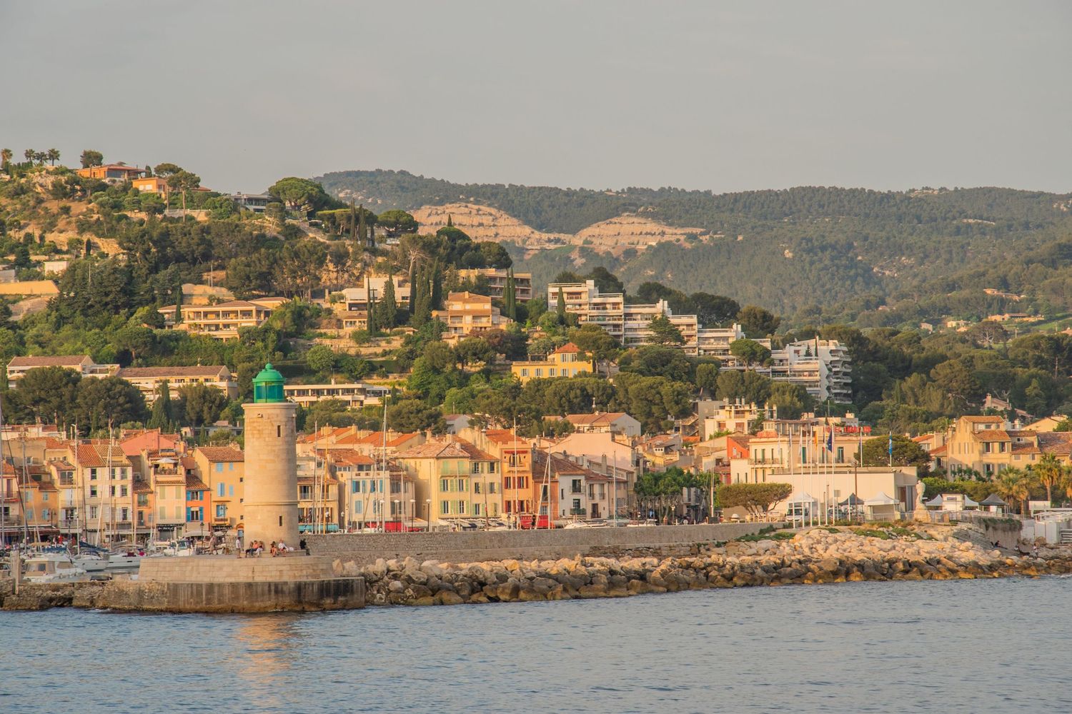 Scenic Mediterranean coastal town with lighthouse and hillside houses overlooking harbor at sunset in French Riviera.