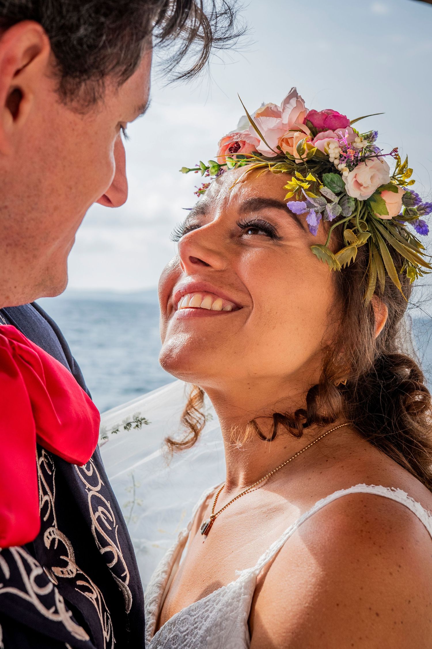 Close up of a bride wearing a colorful floral crown gazing lovingly at her partner against an ocean backdrop.