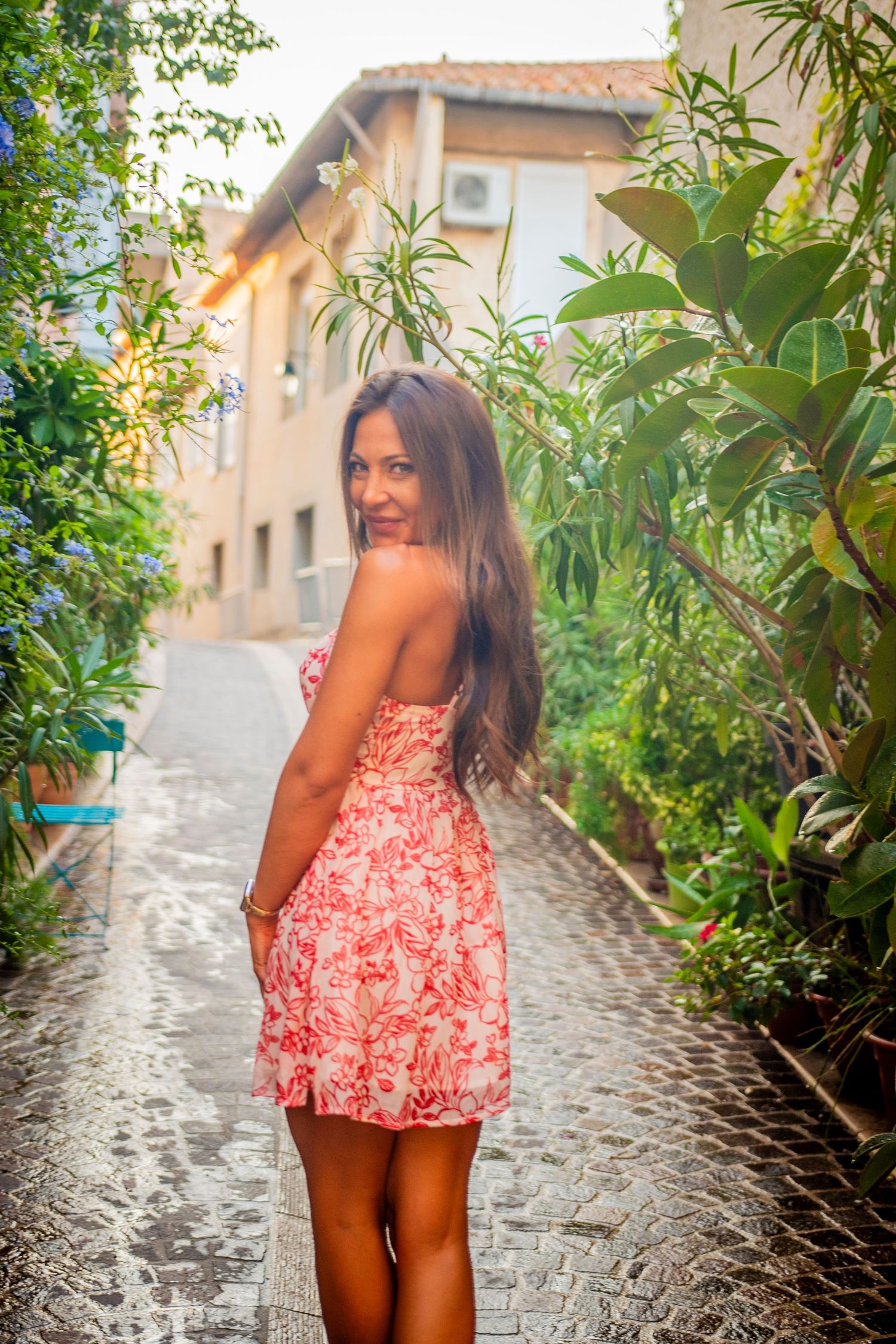 Woman in a floral red dress posing on a cobblestone path surrounded by lush greenery.