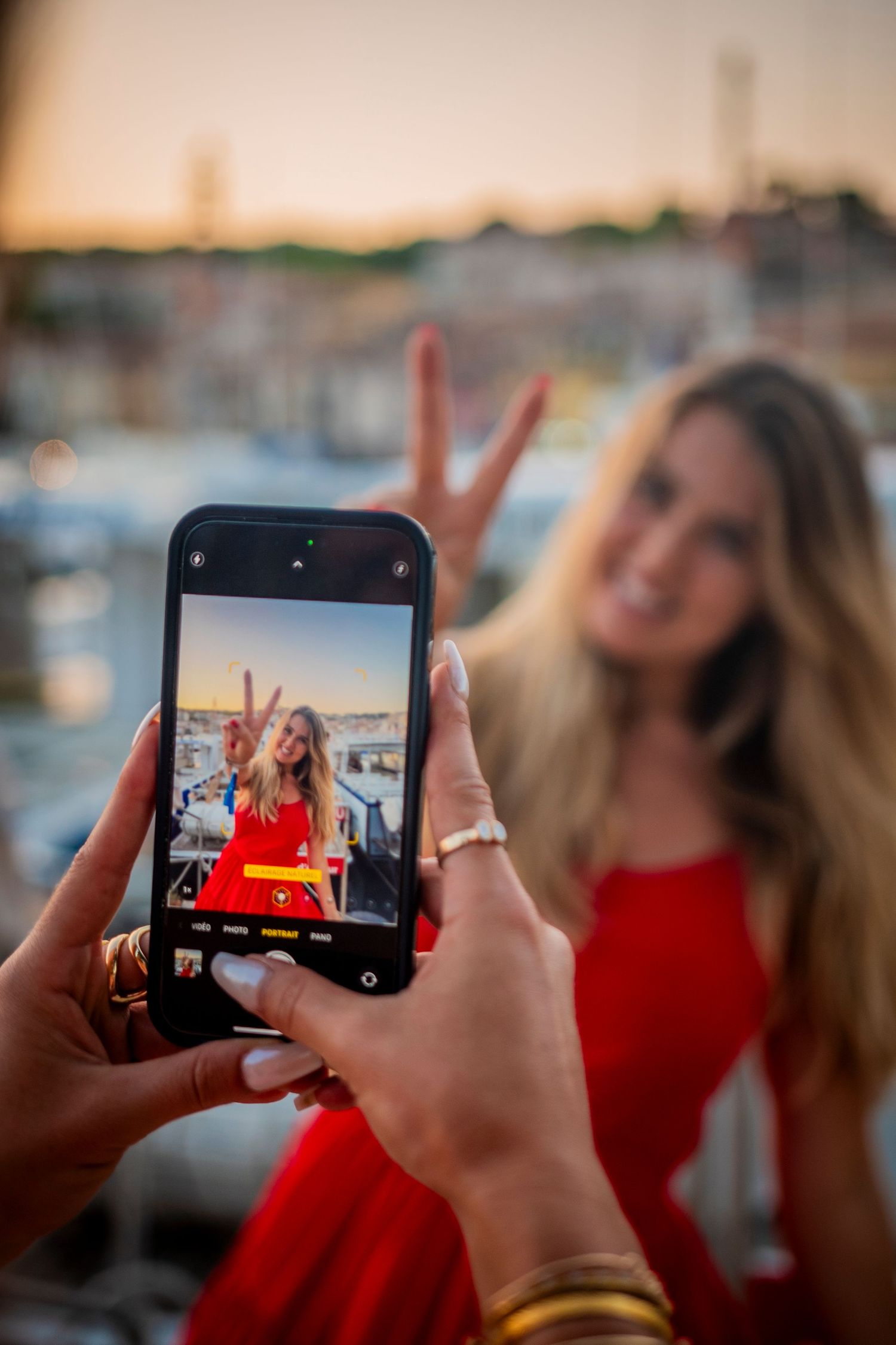 Someone taking a photo on a phone of a person in a red dress making a peace sign against a marina at sunset.