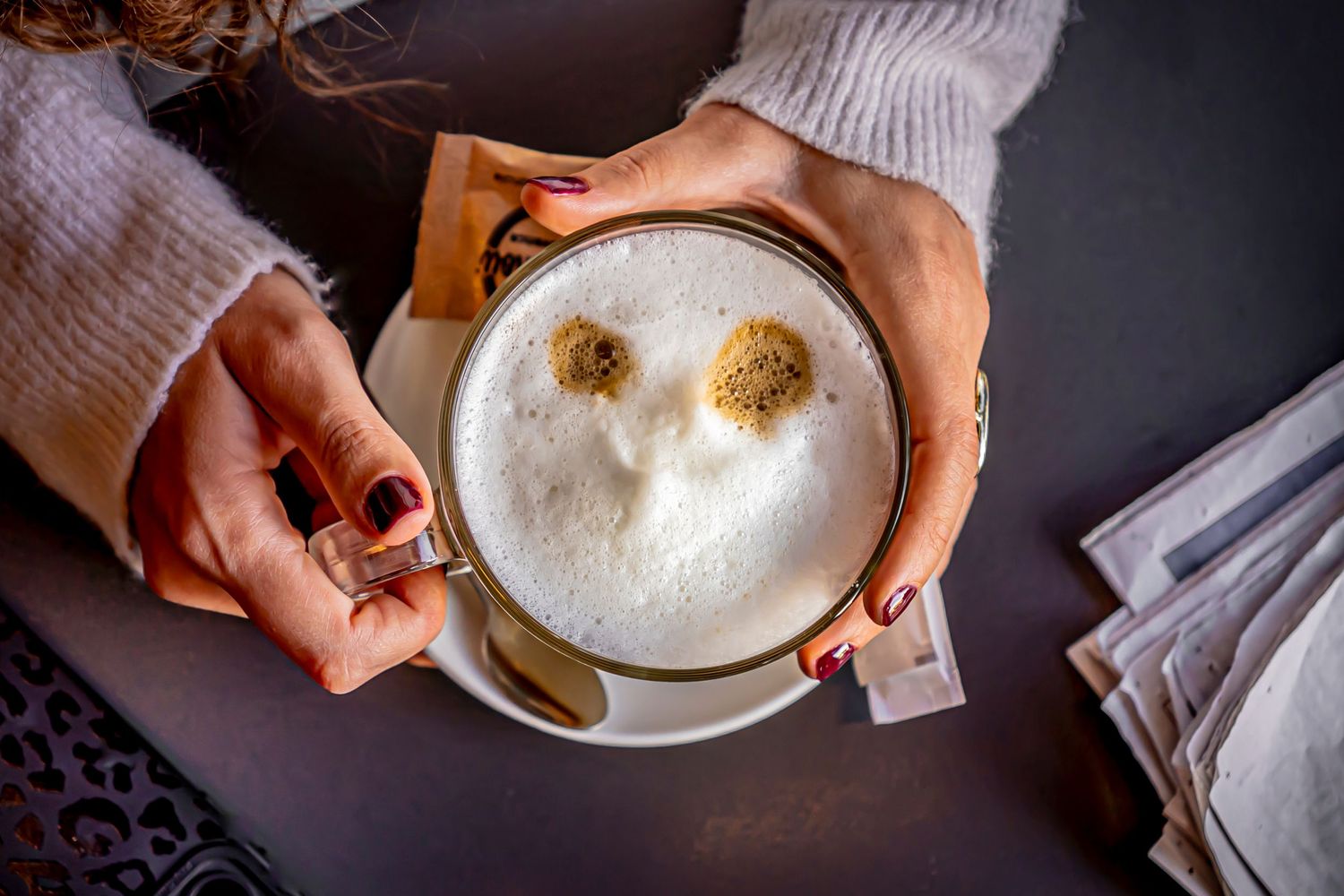 Hands hold coffee cup with frothy latte art in dark moody setting.