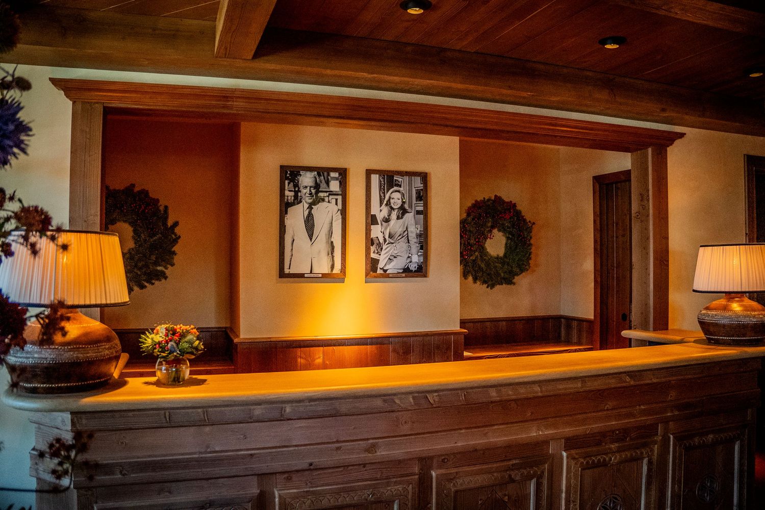 A rustic wooden bar counter with table lamps, framed artwork, and a decorative wreath in warm lighting.
