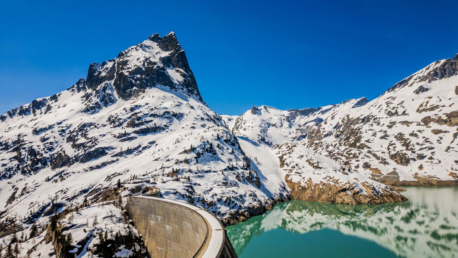 A snow-covered mountain peak reflects in turquoise alpine lake waters with a curved concrete dam wall in the foreground.