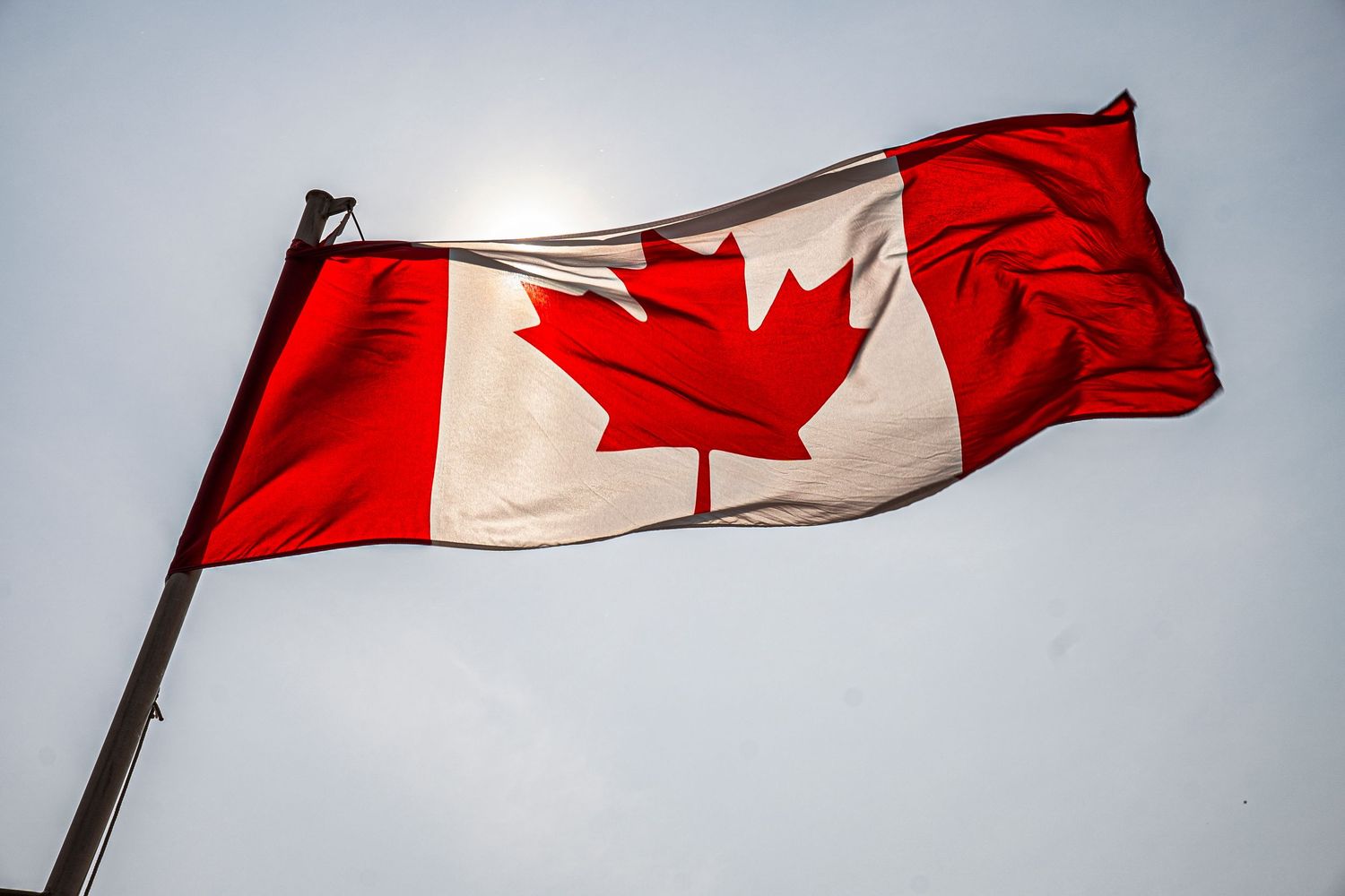 A Canadian flag with red maple leaf emblem waves proudly against a bright cloudy sky.