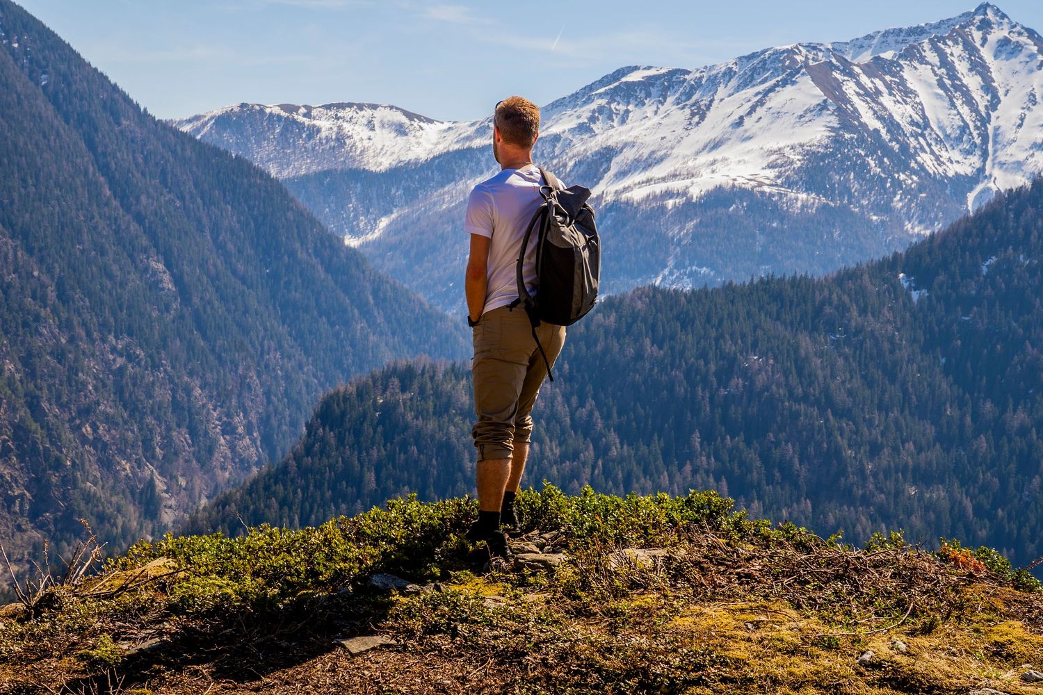 Hiker stands atop a grassy mountain ridge overlooking snow-capped peaks in a dramatic mountain landscape.