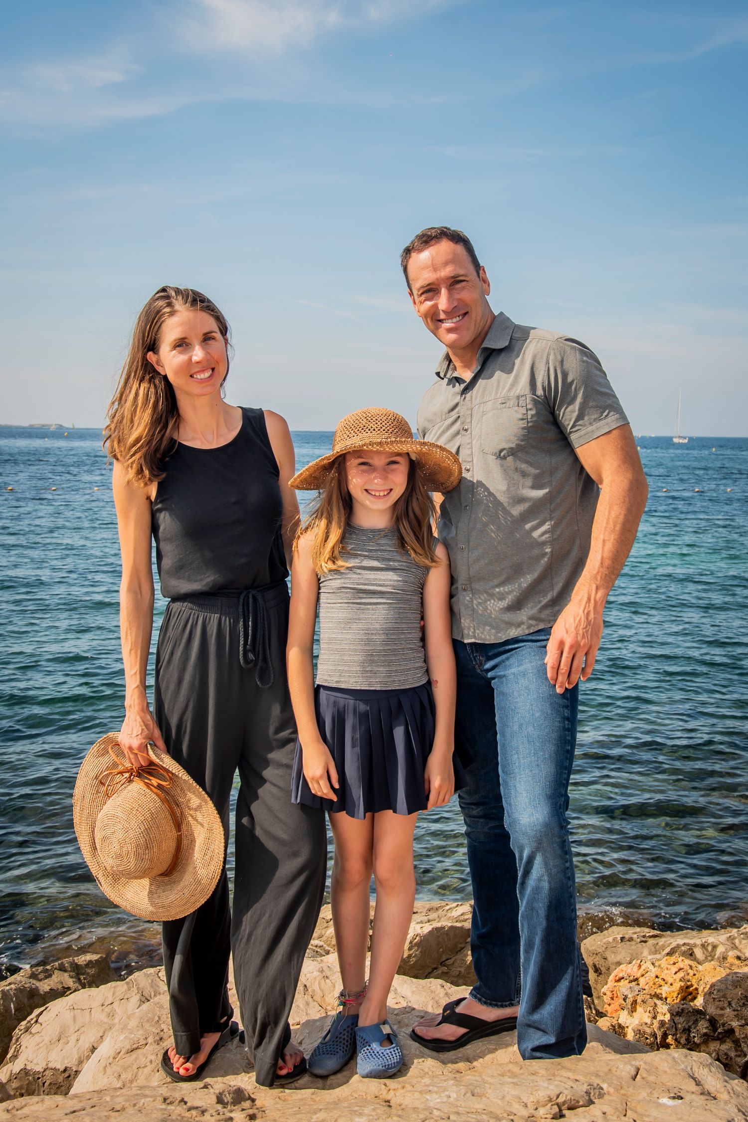 Family standing together on rocky coastline with bright blue ocean waters in background.