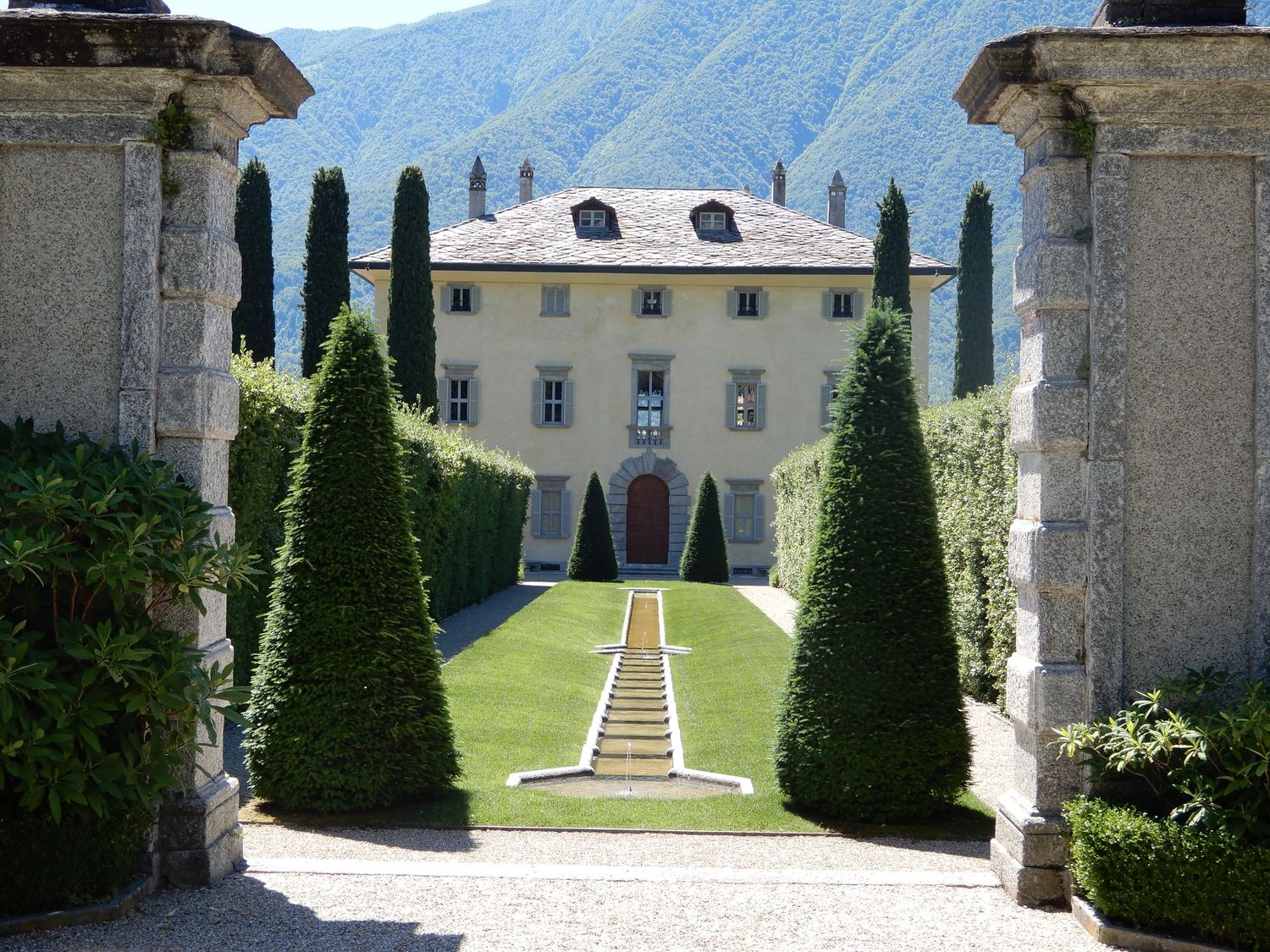 Elegant Italian villa with manicured topiary gardens, stone pillars, and cypress trees framing a grand staircase entrance.