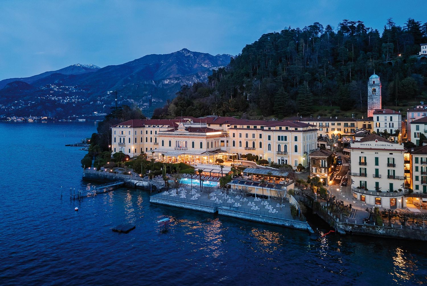 Aerial dusk view of a luxury lakeside hotel on Lake Como, Italy, with mountain backdrop and illuminated waterfront.