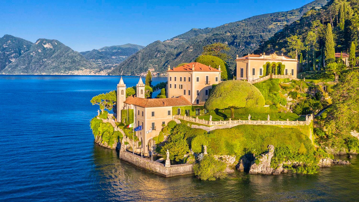 Aerial view of Villa del Balbianello on Lake Como, Italy, surrounded by lush greenery and crystal-clear blue waters.