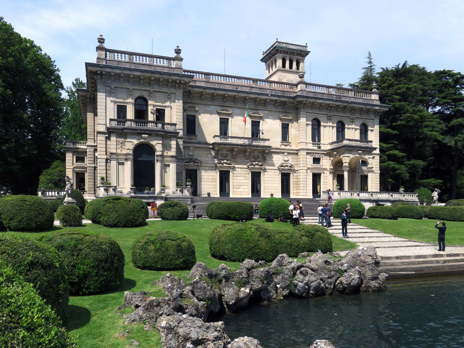 Elegant Italian villa with manicured hedges, rocky shoreline, and visitors strolling the lakeside gardens on a cloudy day.