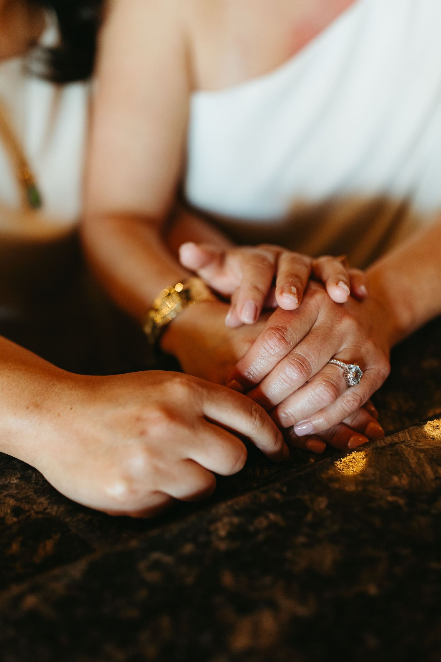 Close-up of bride and groom holding hands at wedding, showcasing diamond engagement ring and gold bracelet.