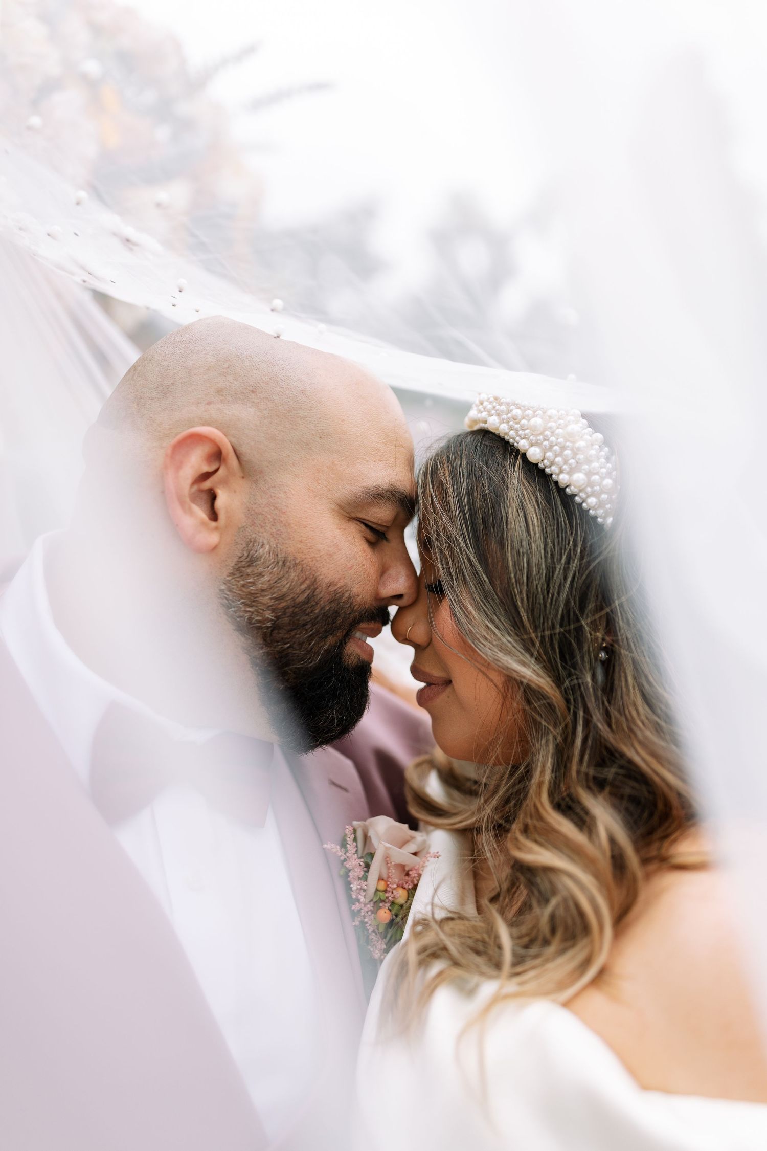 Romantic close-up of bride and groom touching foreheads under a flowing veil, bride wearing a pearl crown headpiece.
