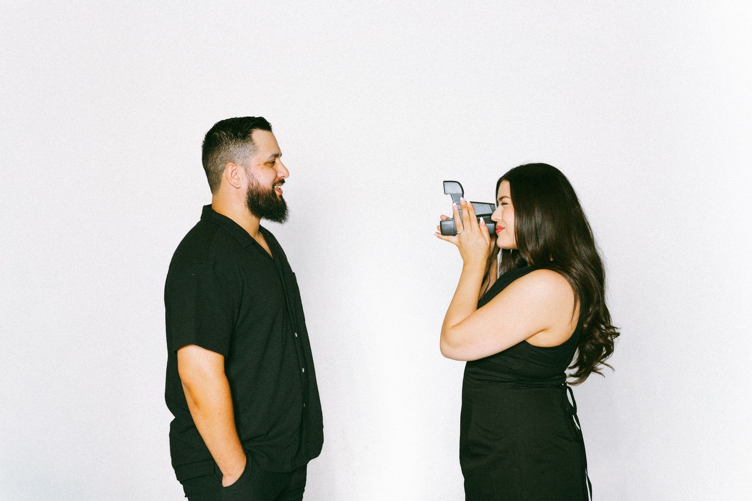 Engagement studio session. A woman in black dress photographs a bearded man in black shirt against a white background.