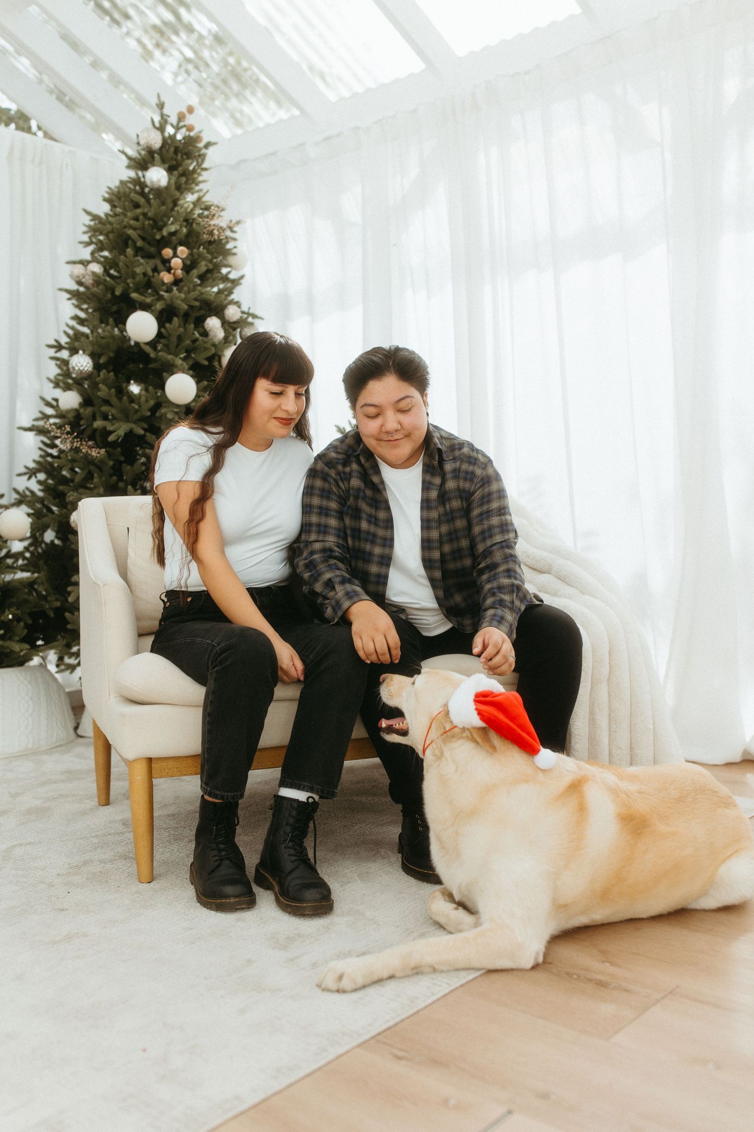 Couple sitting on sofa petting a Labrador wearing a Santa hat near a decorated Christmas tree.