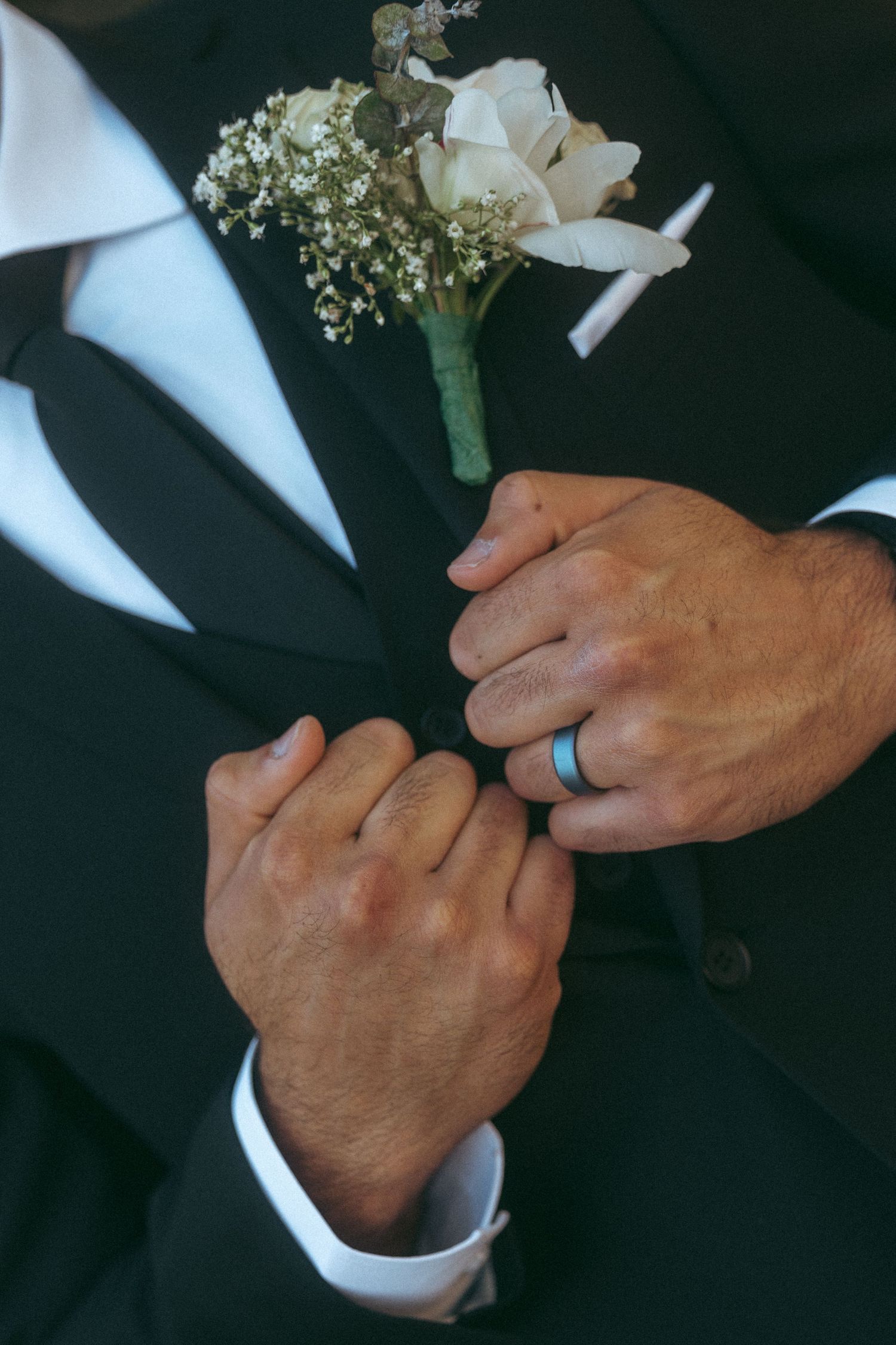 Groom in black suit adjusts white rose boutonniere, wearing a blue wedding band on his finger.