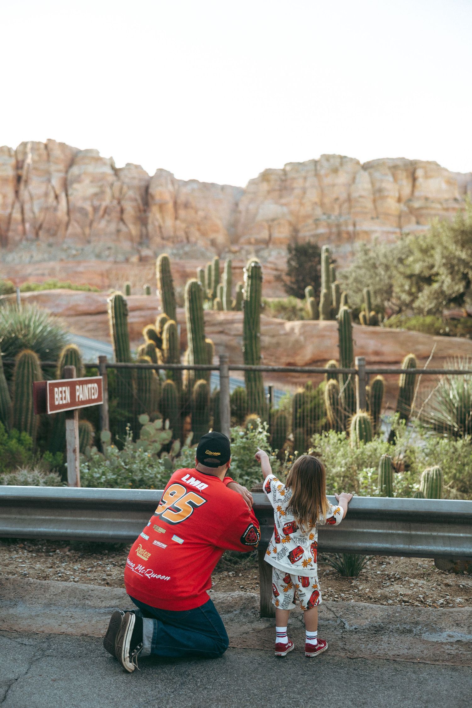 A father and child in Lightning McQueen jerseys point at cacti and red rock formations at Cars Land, Disney California Adventure.