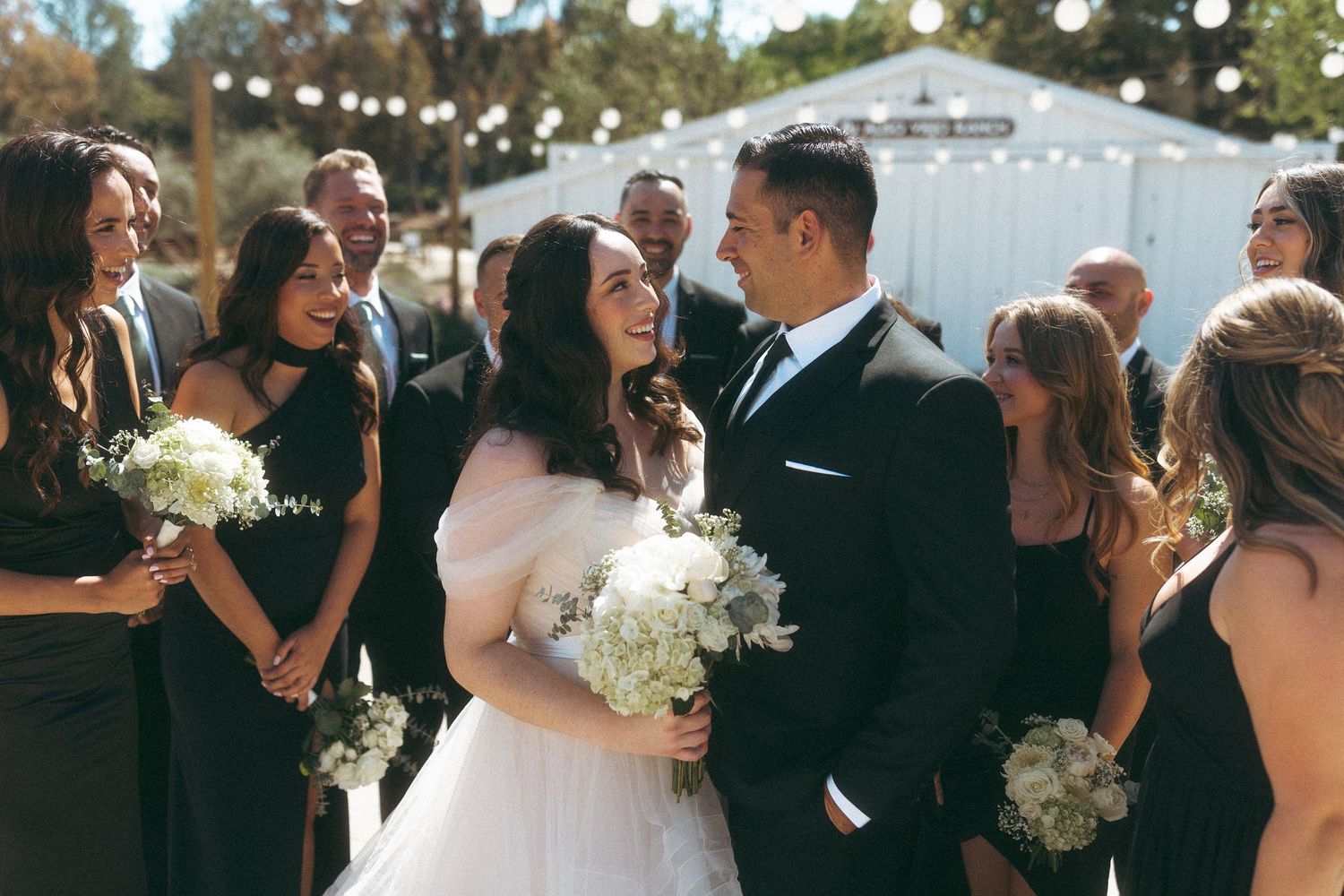 Bride and groom laughing joyfully surrounded by wedding party in black attire with white floral bouquets.