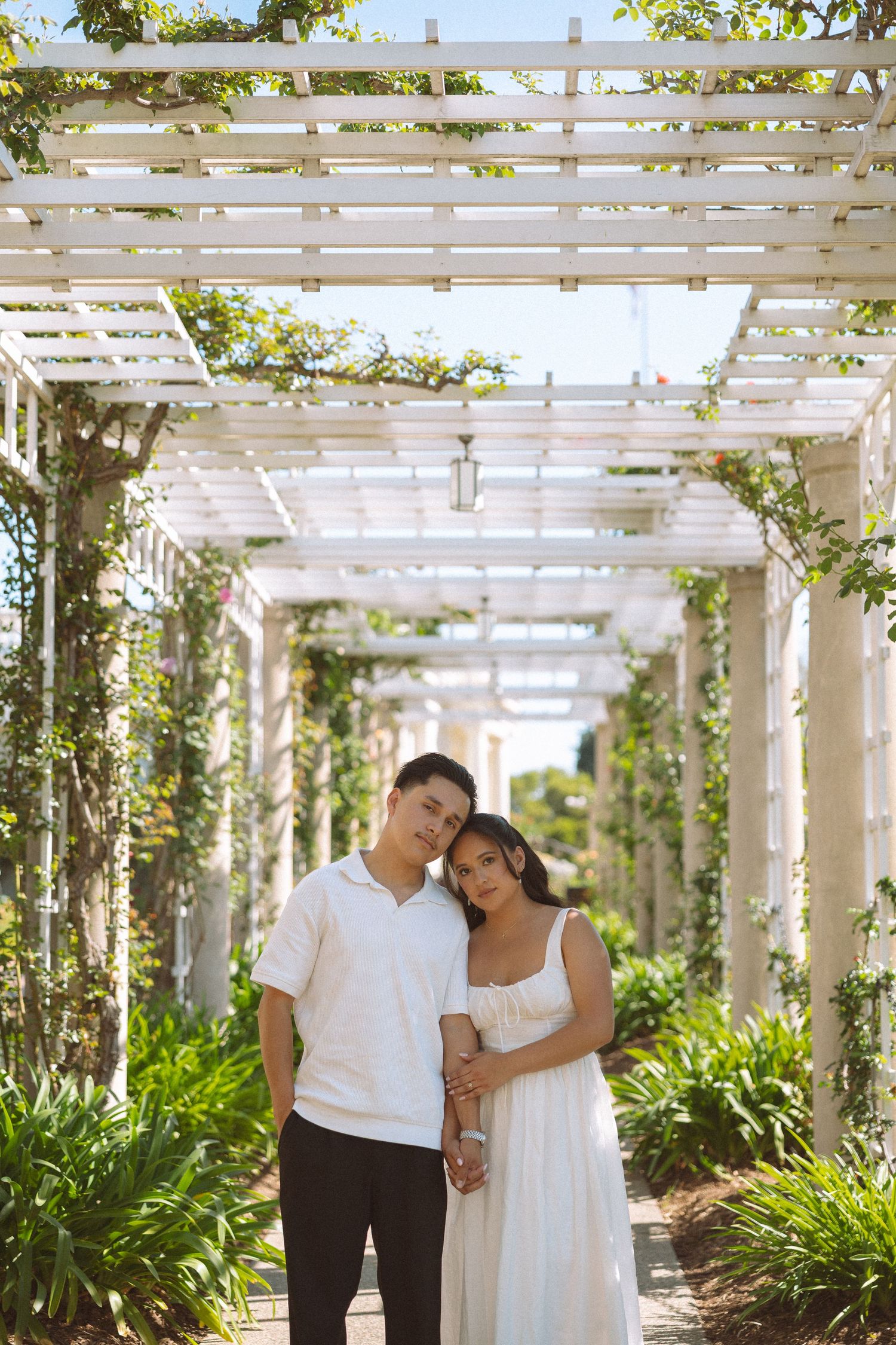 A couple poses under a white pergola covered in greenery, the woman in a white dress leaning on the man. Bridgerton theme engagement session
