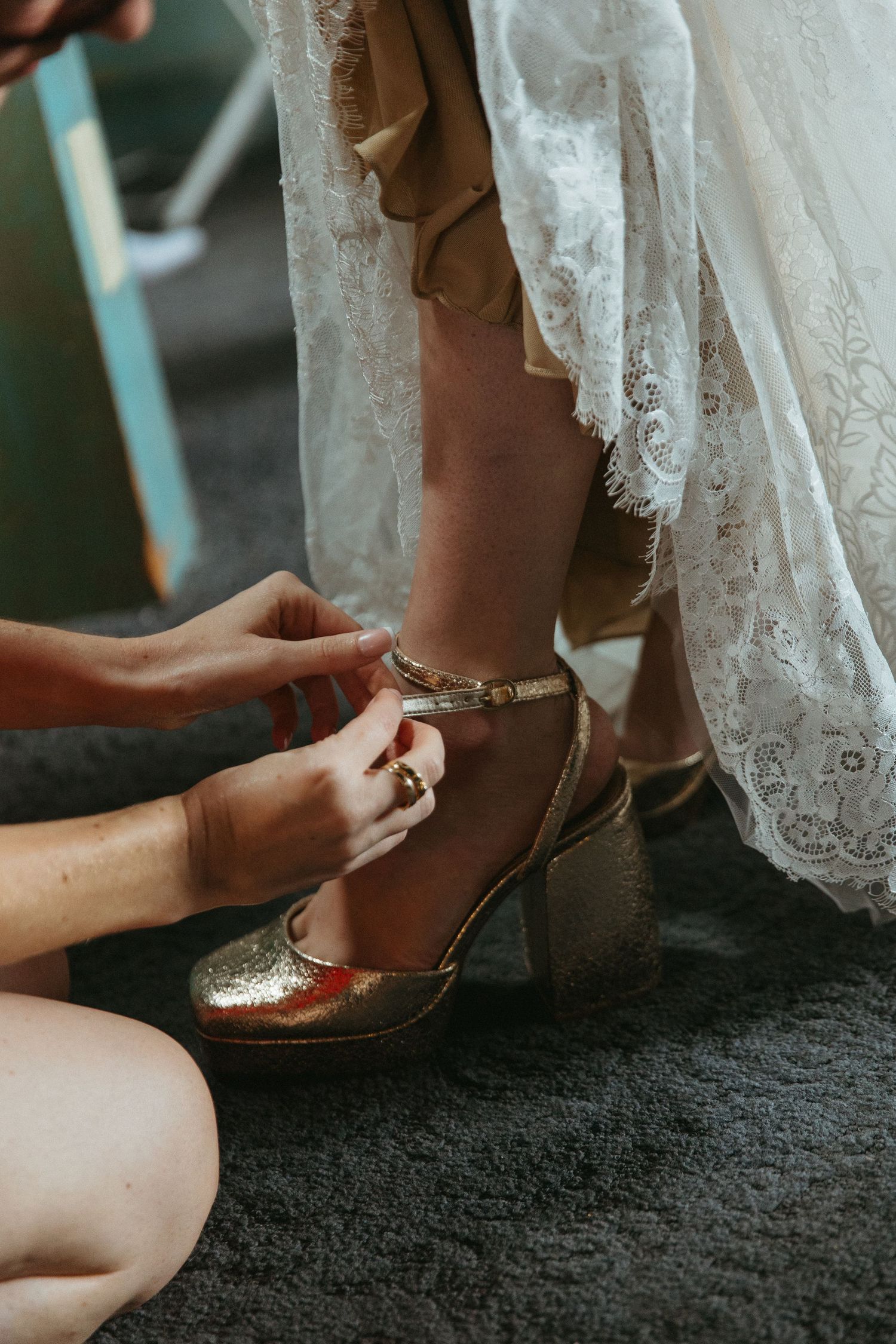 Bridesmaid buckles gold block-heel wedding shoes on bride wearing lace dress during bridal preparations.