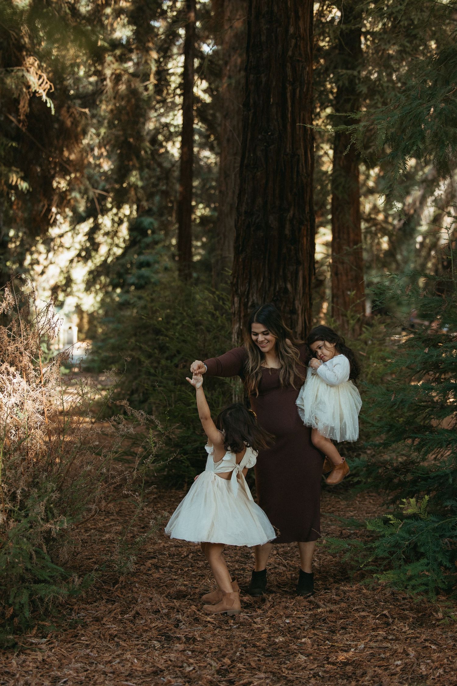 A mother in a brown dress holds one toddler while dancing with another in white dresses among tall redwood trees.