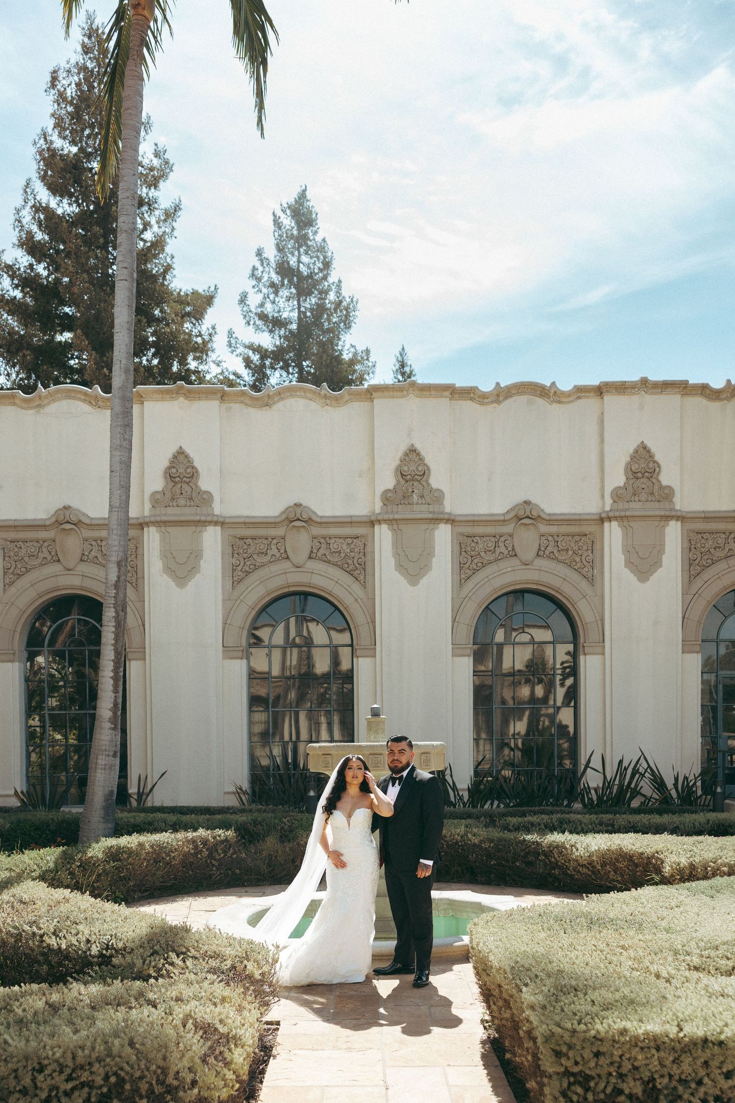 Bride and groom posing in front of an ornate white building with manicured hedges.