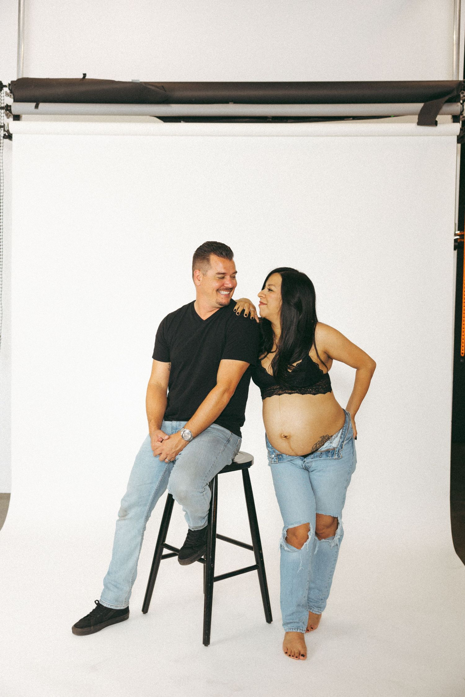 Couple in a photography studio, pregnant woman in jeans and bralette with partner on stool, sharing a joyful moment.