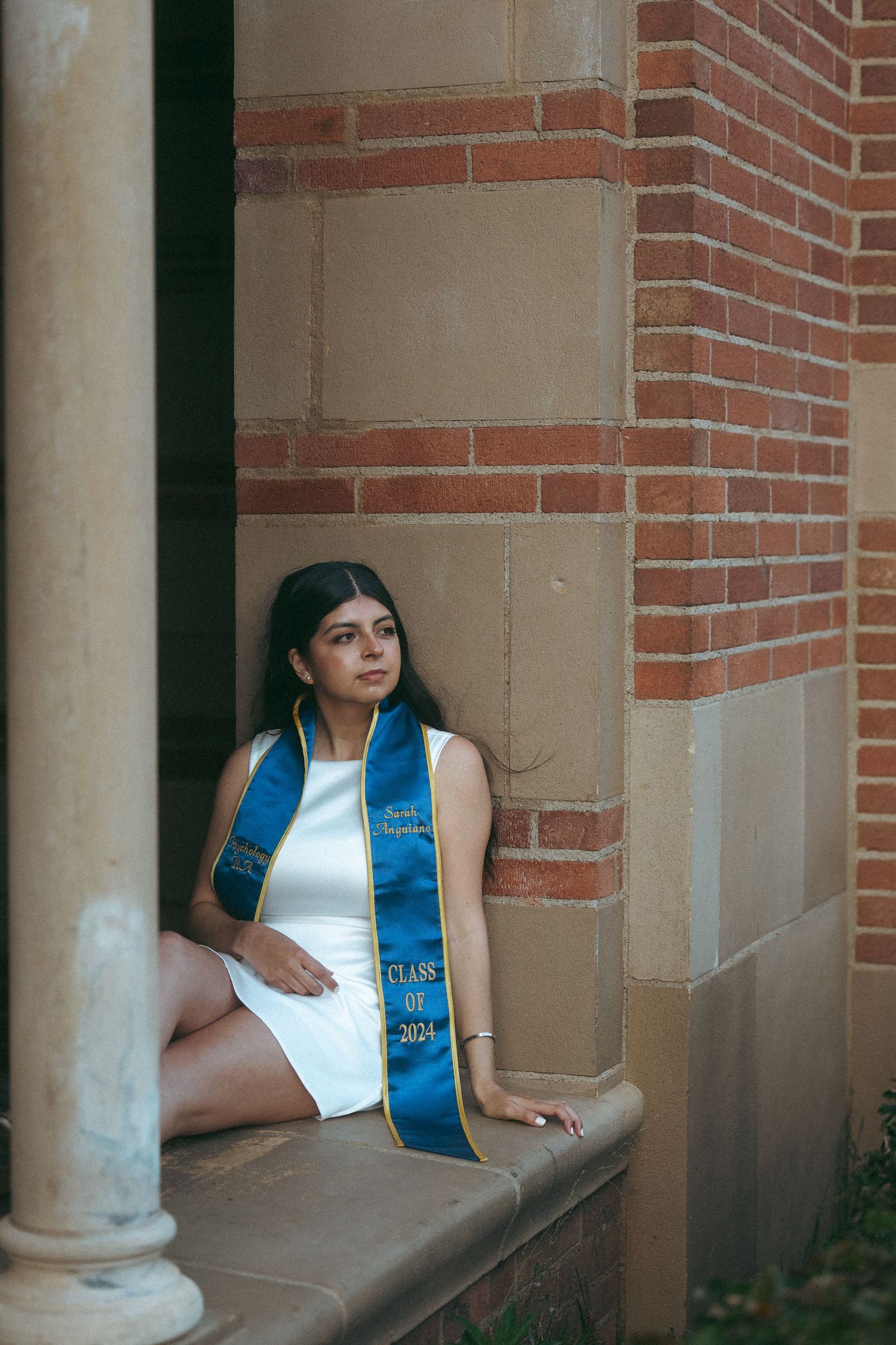 A young woman in a white dress and blue graduation stole sits on stone steps beside a brick wall, Class of 2024.