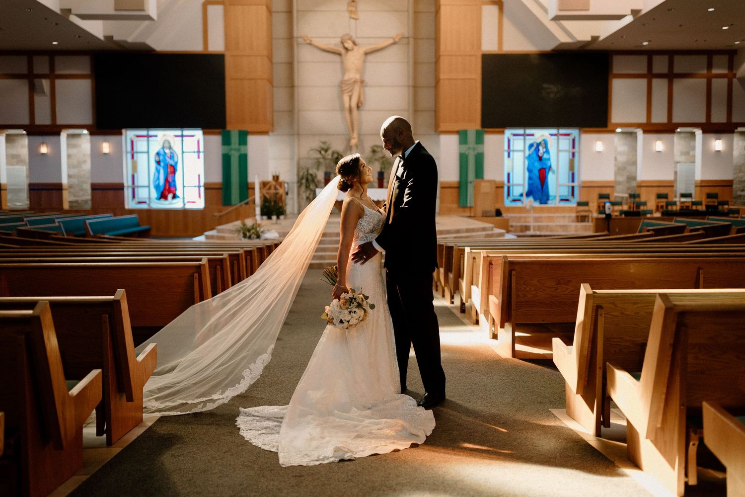 Bride and groom stand facing each other in a stunning church aisle, with a crucifix and stained glass windows behind them.
