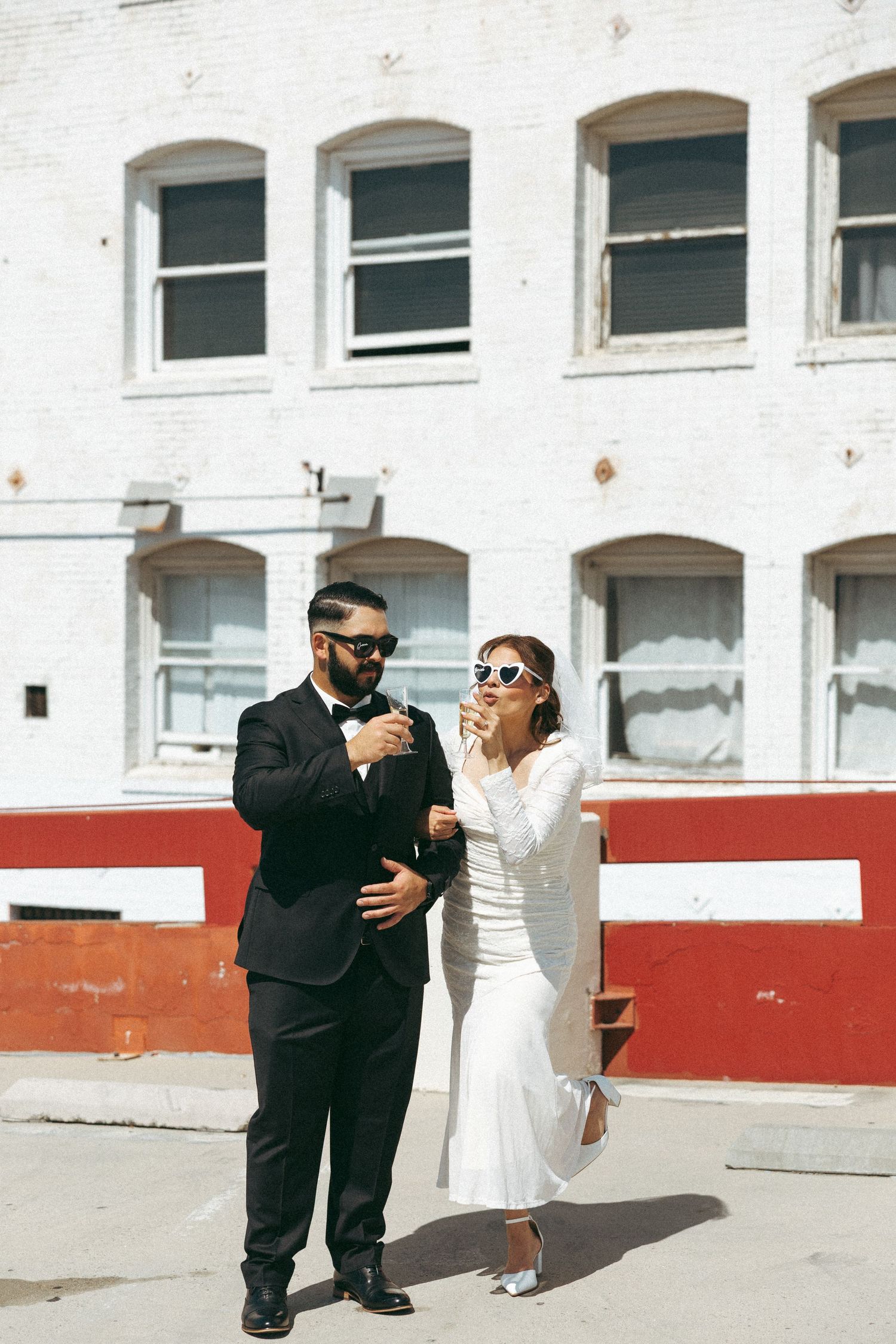 Bride and groom in sunglasses celebrate outside a white brick building, sharing champagne in stylish wedding attire.
