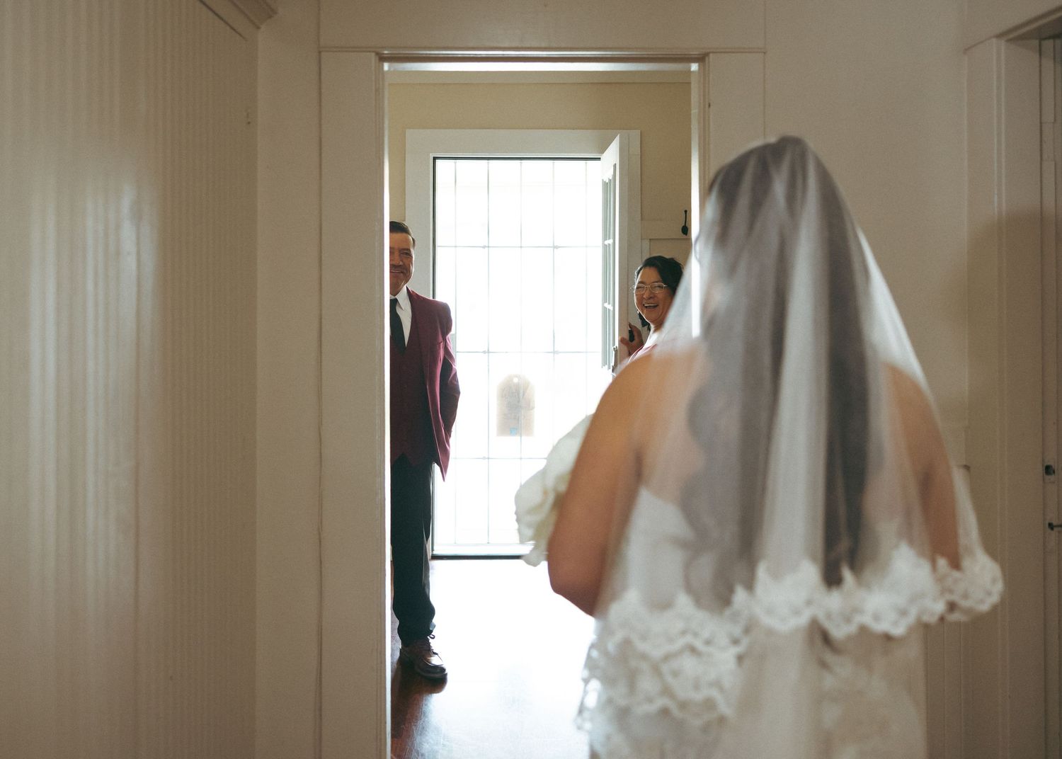 Bride in lace veil walks toward open doorway where guests await during wedding day preparations.