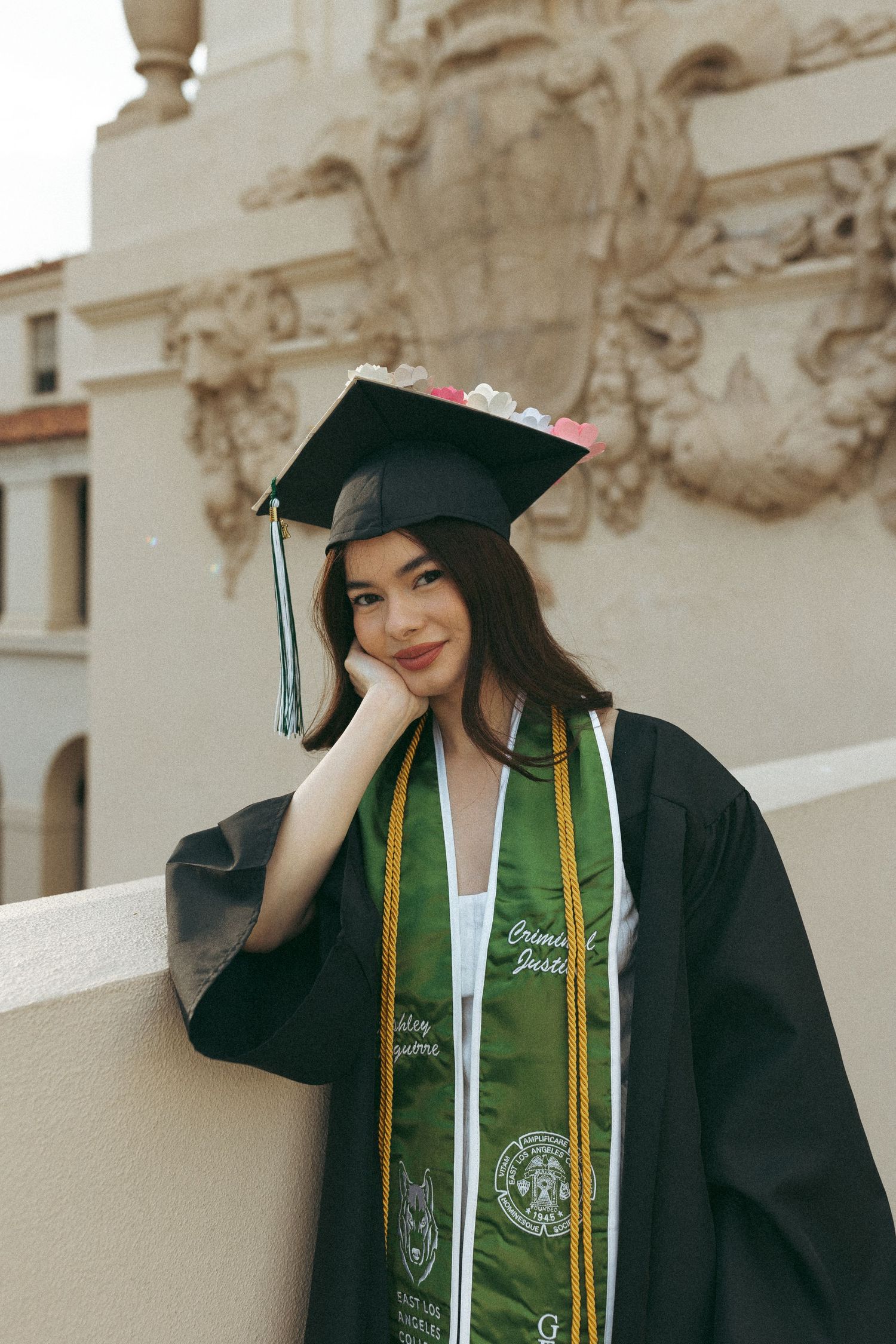 A young woman in graduation cap and gown with green honor stoles poses at an ornate building.